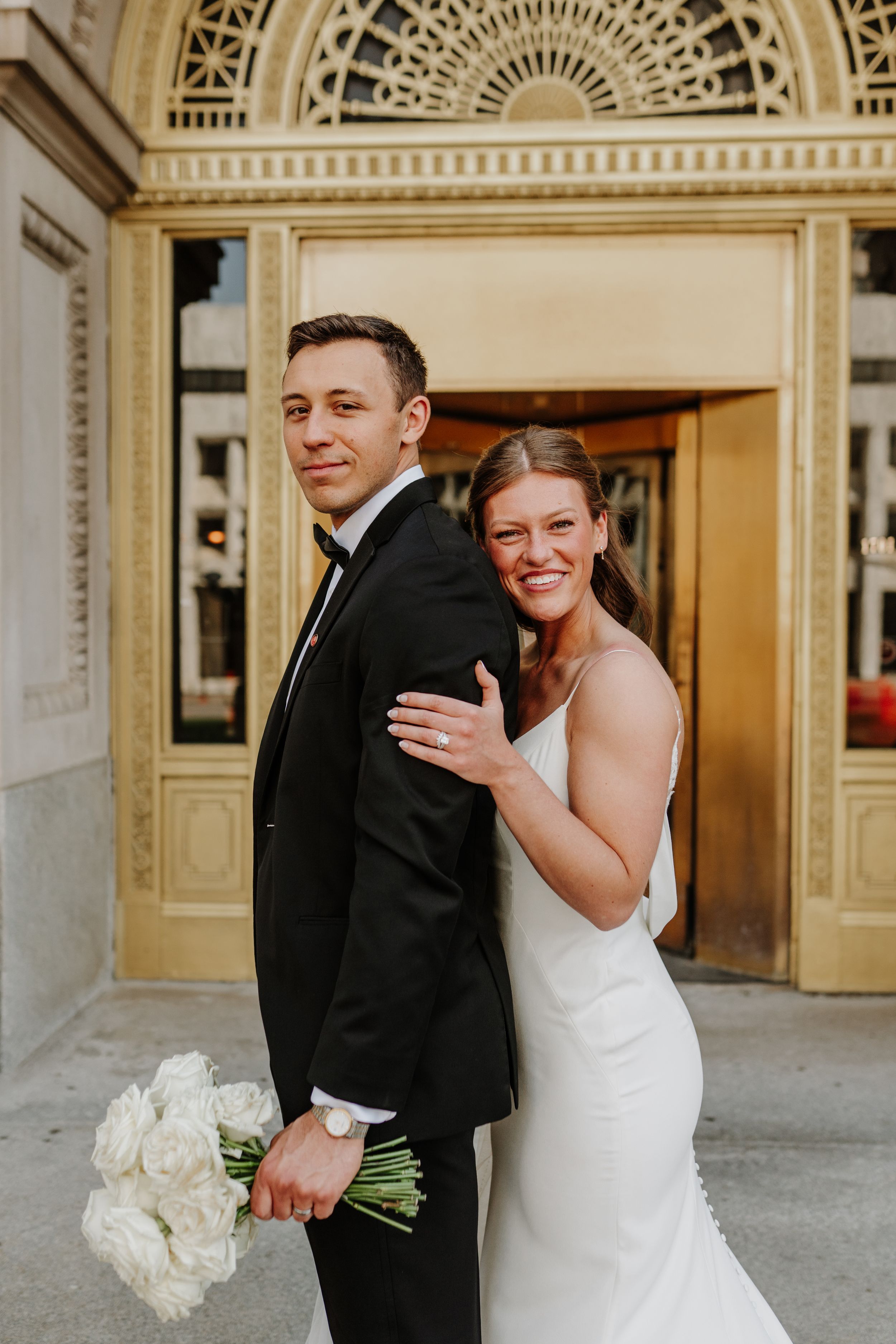 Joyful wedding couple poses outside ornate building entrance with white bouquet.