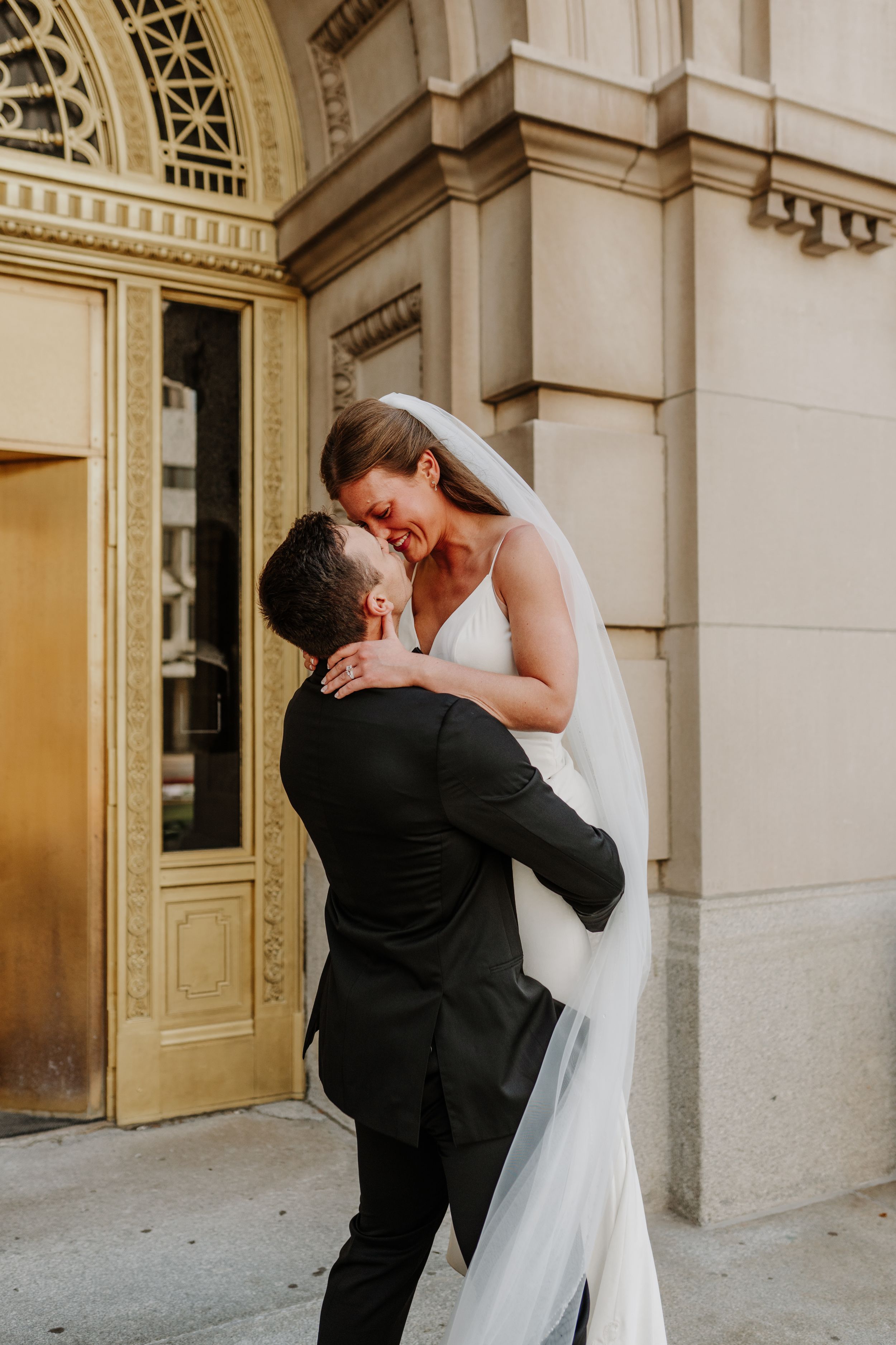 Groom lifts bride in romantic embrace outside historic building with golden doorway.