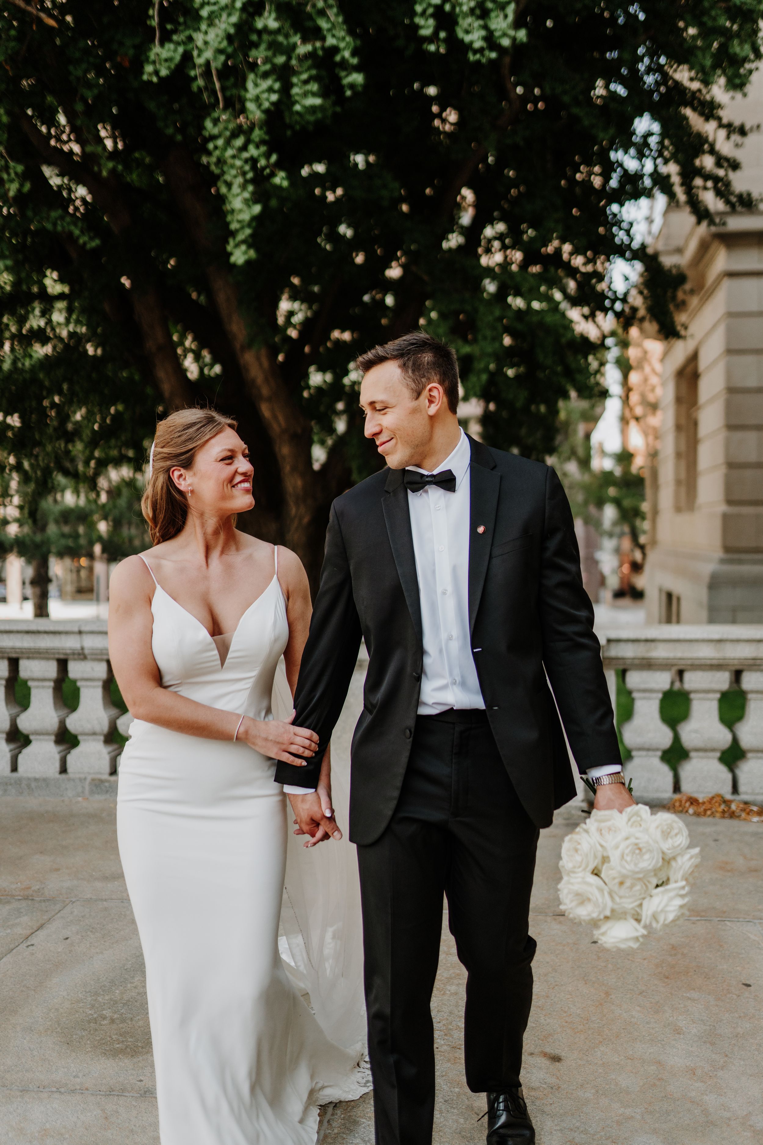 Bride in fitted white gown and groom in tuxedo walking outdoors near stone balustrade.