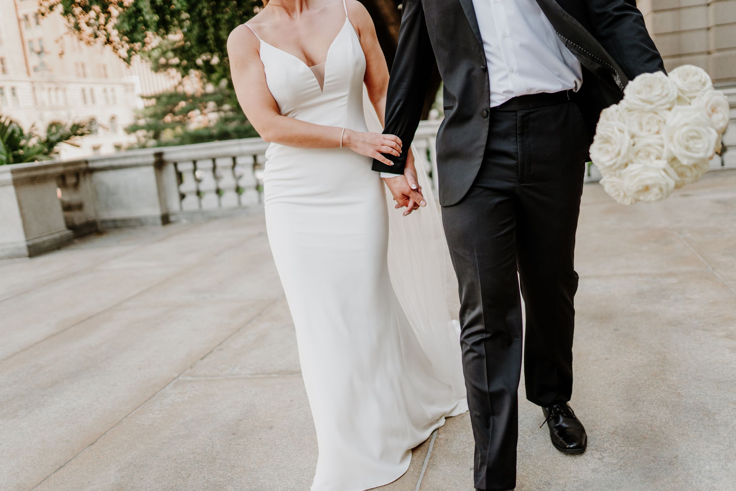 Elegant wedding couple holding hands while walking in formal attire with white floral bouquet.