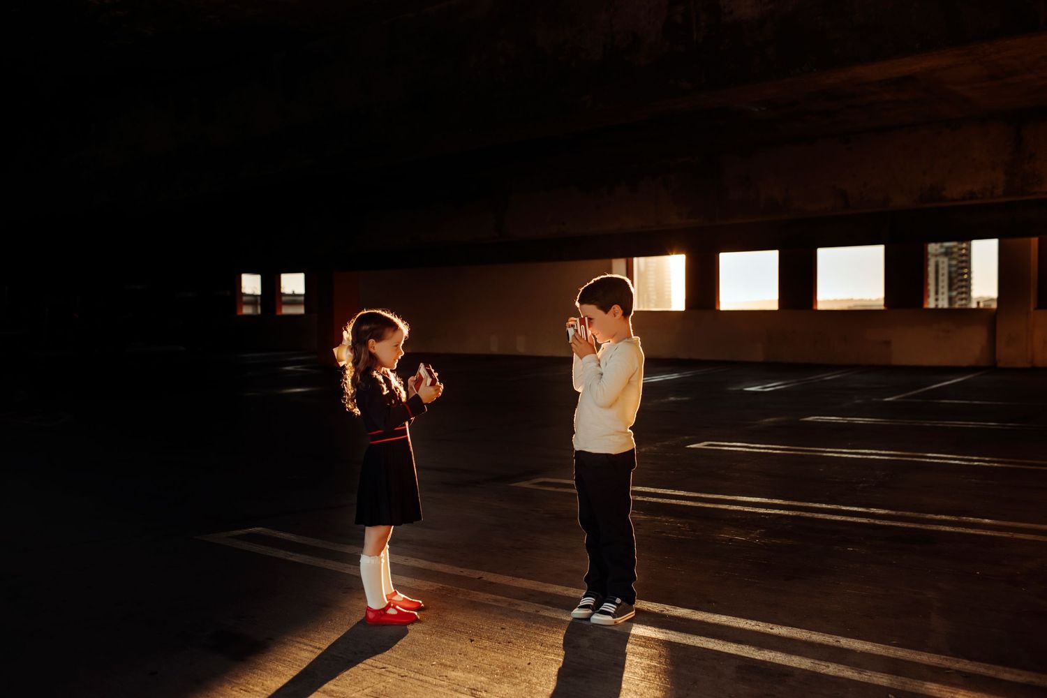 Two kids holding retro cameras, playfully pointing them at each other in an urban parking structure, creating a fun and nostalgic vibe.