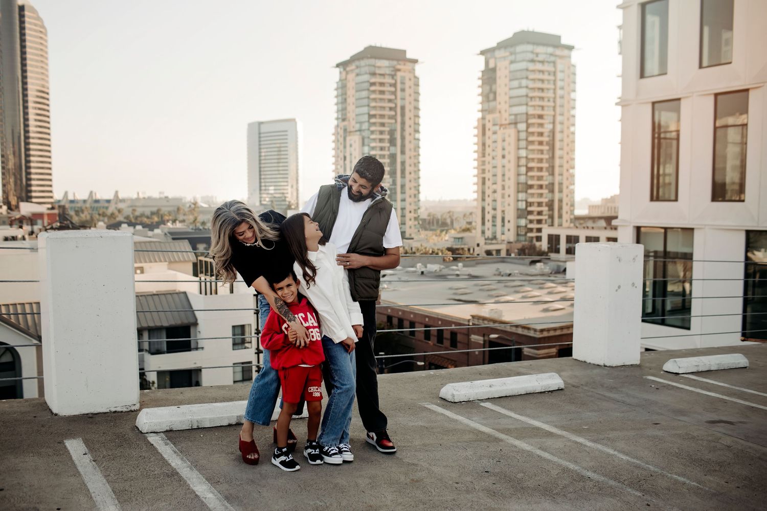 A family of four posing casually on a rooftop parking lot, dressed in relaxed outfits, sharing smiles against an urban backdrop.