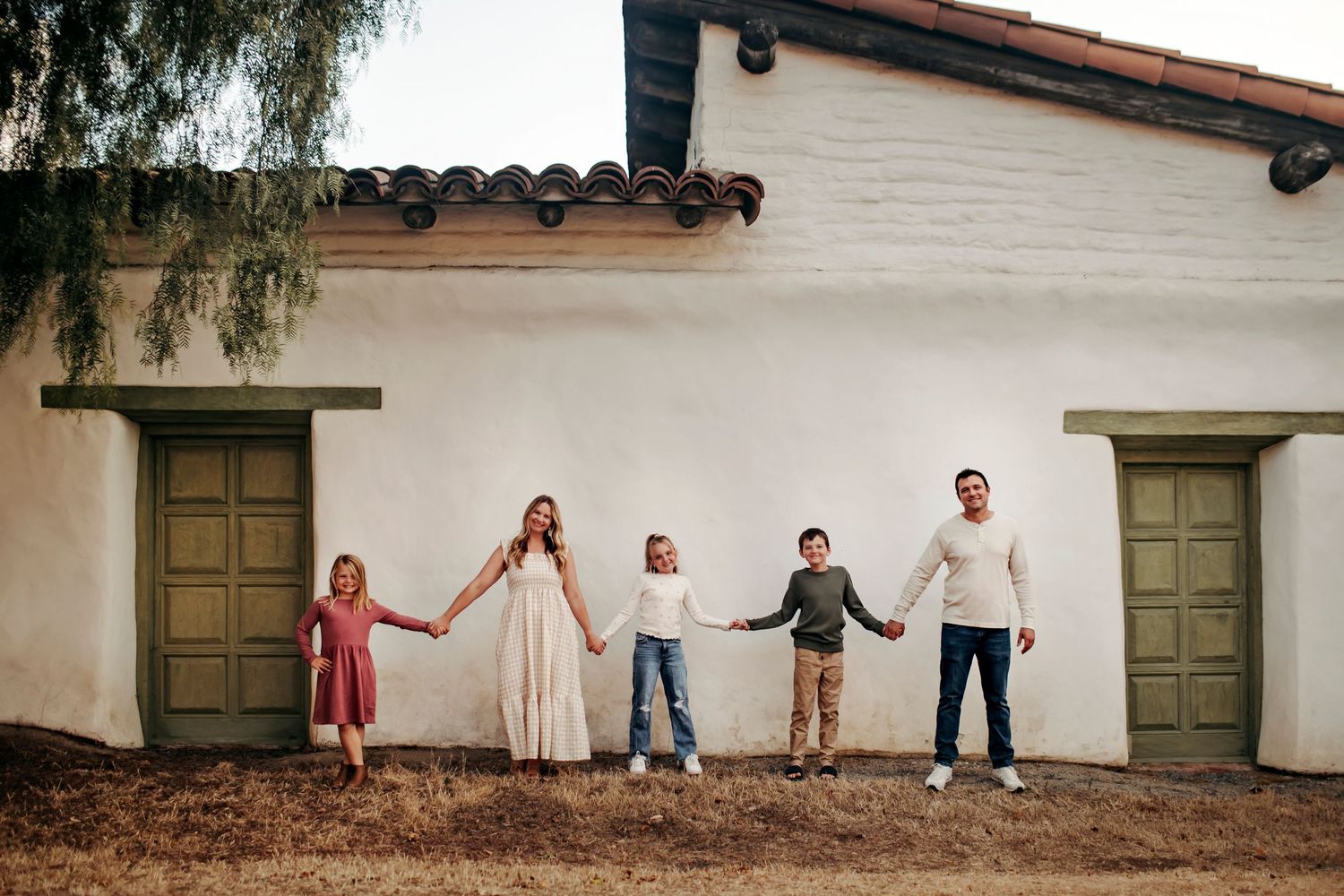 A family of five holding hands in a line, walking together in Old Town San Diego, surrounded by historic buildings and a cheerful atmosphere
