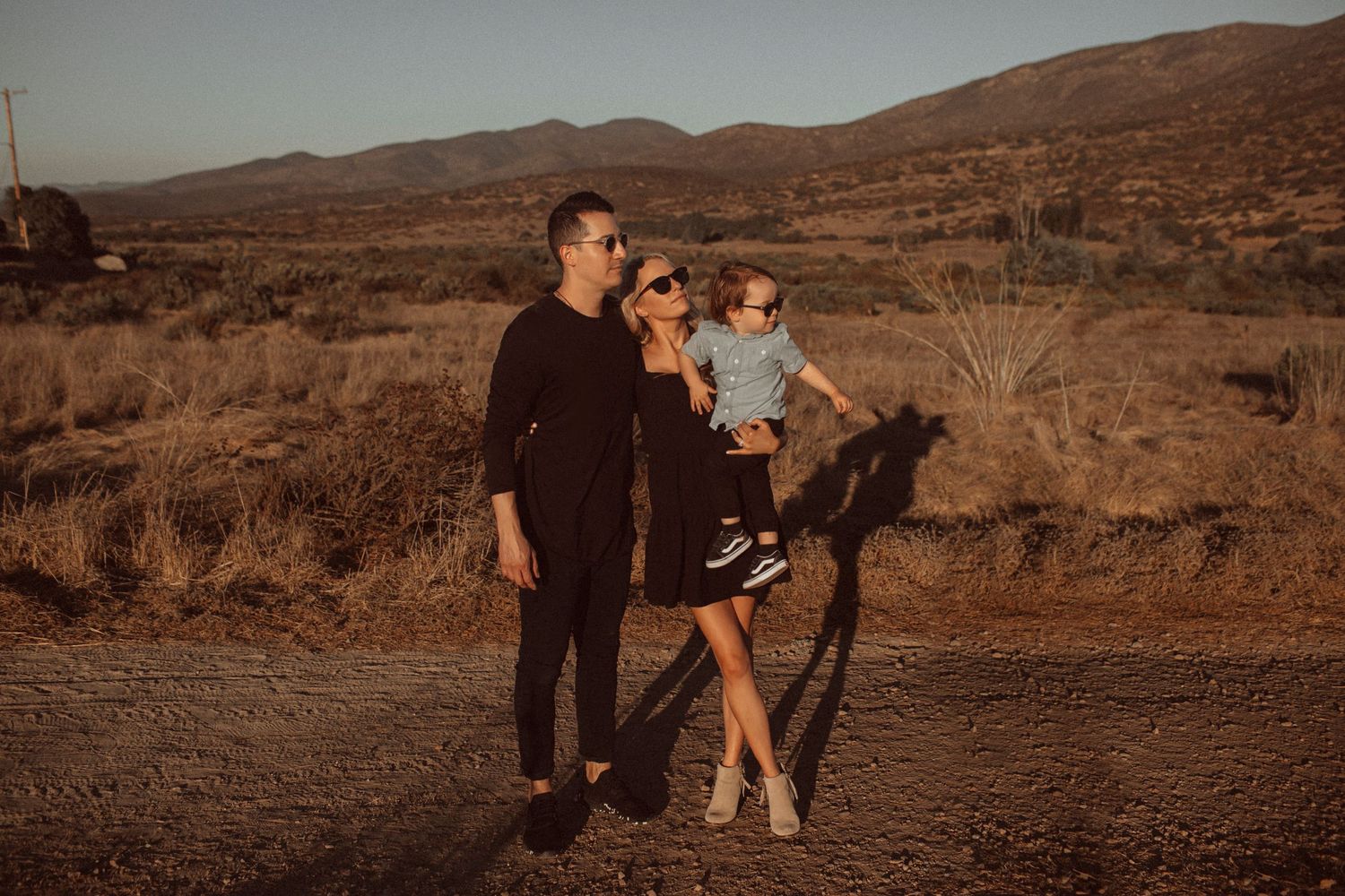 A mom and dad holding their toddler in a field with mountains in the background, standing in direct light, looking to the side with serious