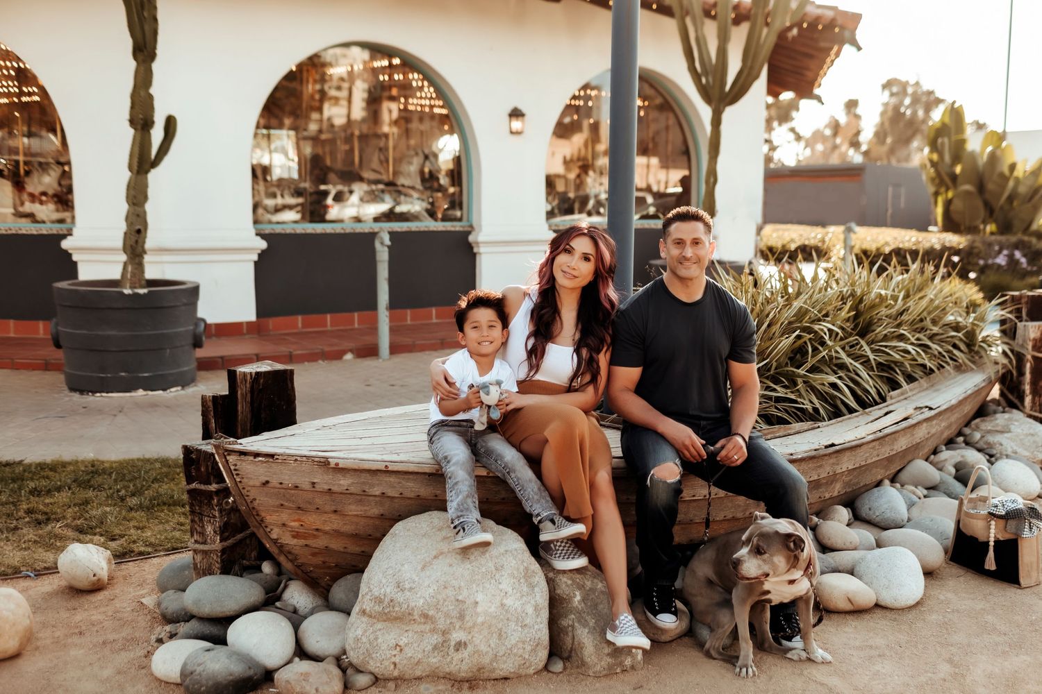 A family of three sitting casually on a ship-themed seat in front of a carousel, sharing smiles and enjoying a fun, playful moment.