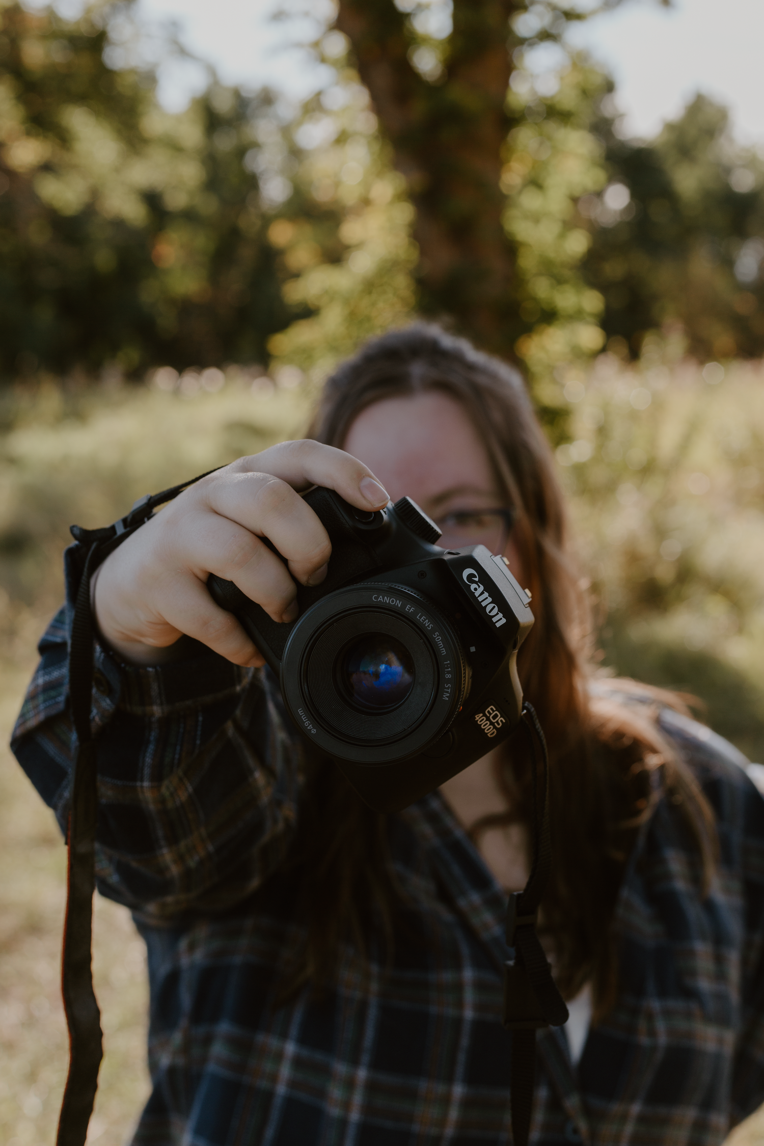 Photographer standing outside in front of a tree holding her camera out in front of her covering her face.
