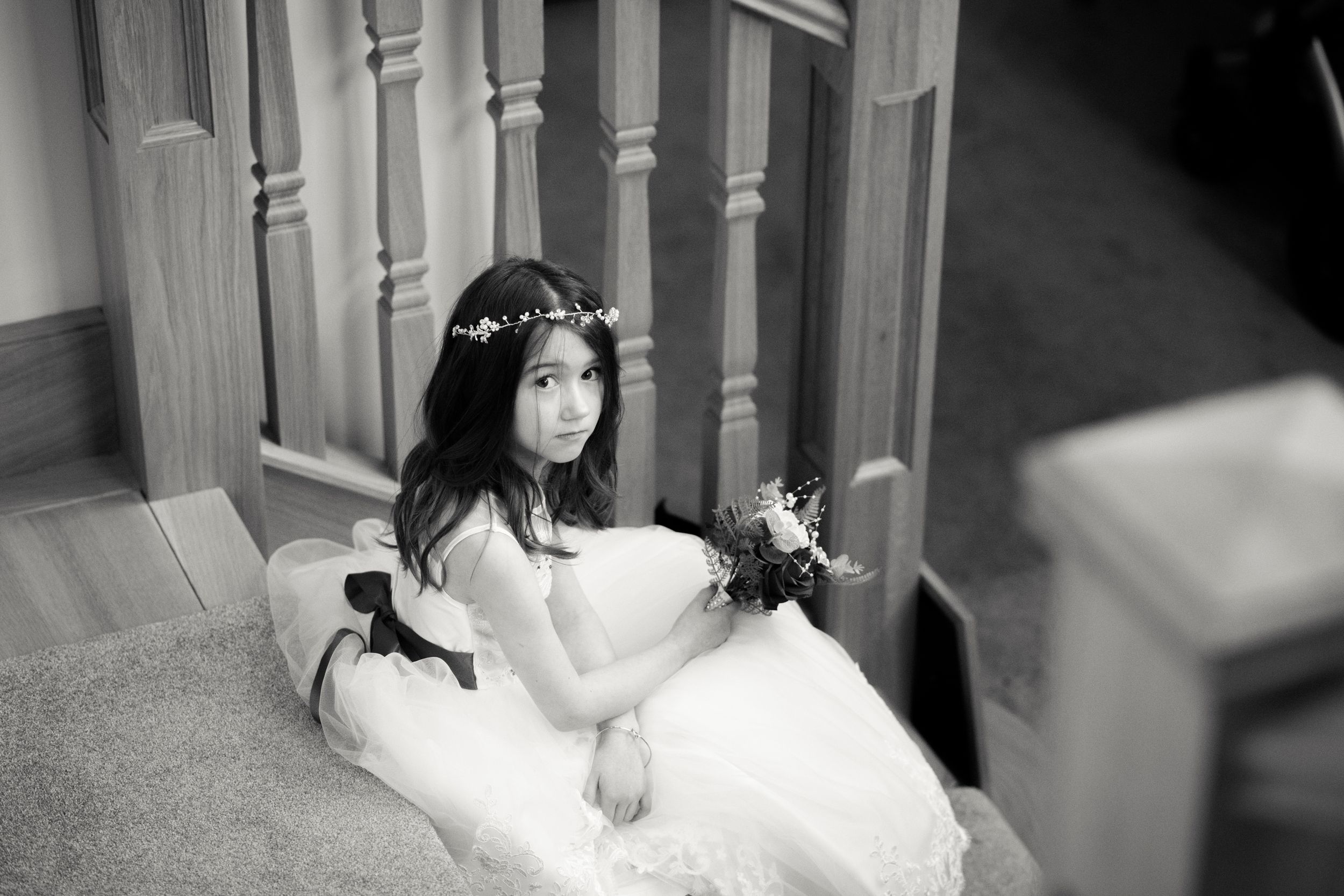 A bride in white dress sits elegantly on carpeted stairs wearing a floral crown.