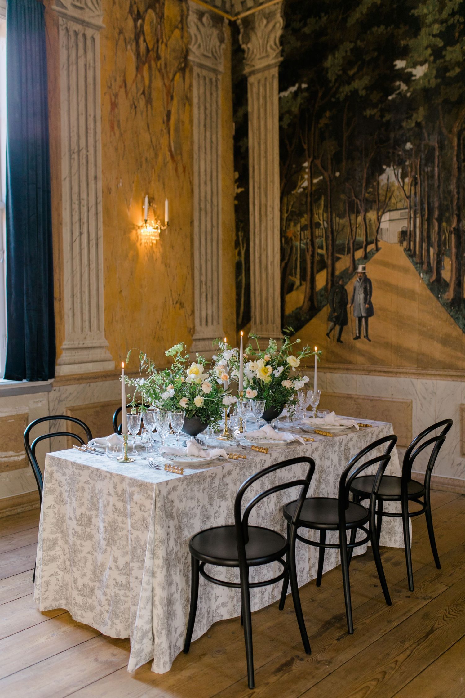 Elegant dining table with white lace tablecloth, black bistro chairs, and floral centerpiece against mural-adorned wall.