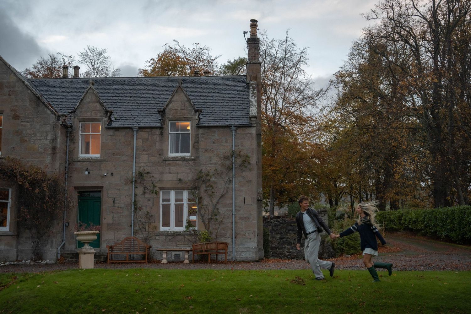 Fashion-forward couple running hand in hand in front of a castle-like house in Scotland, with dreamy fall vibes and rich earthy tones.
