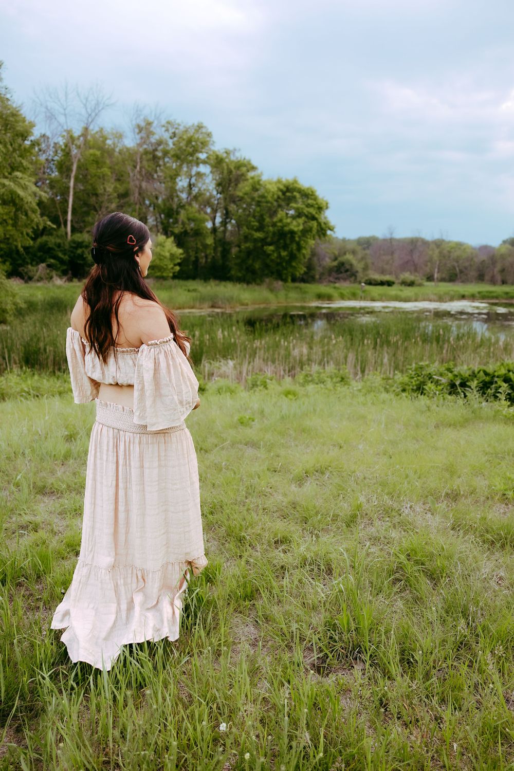 A woman in a flowy white dress stands in a serene meadow overlooking a tranquil pond on a spring day.