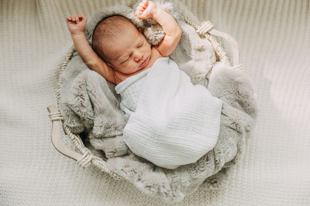Newborn baby wrapped in white stretching against soft gray blanket.