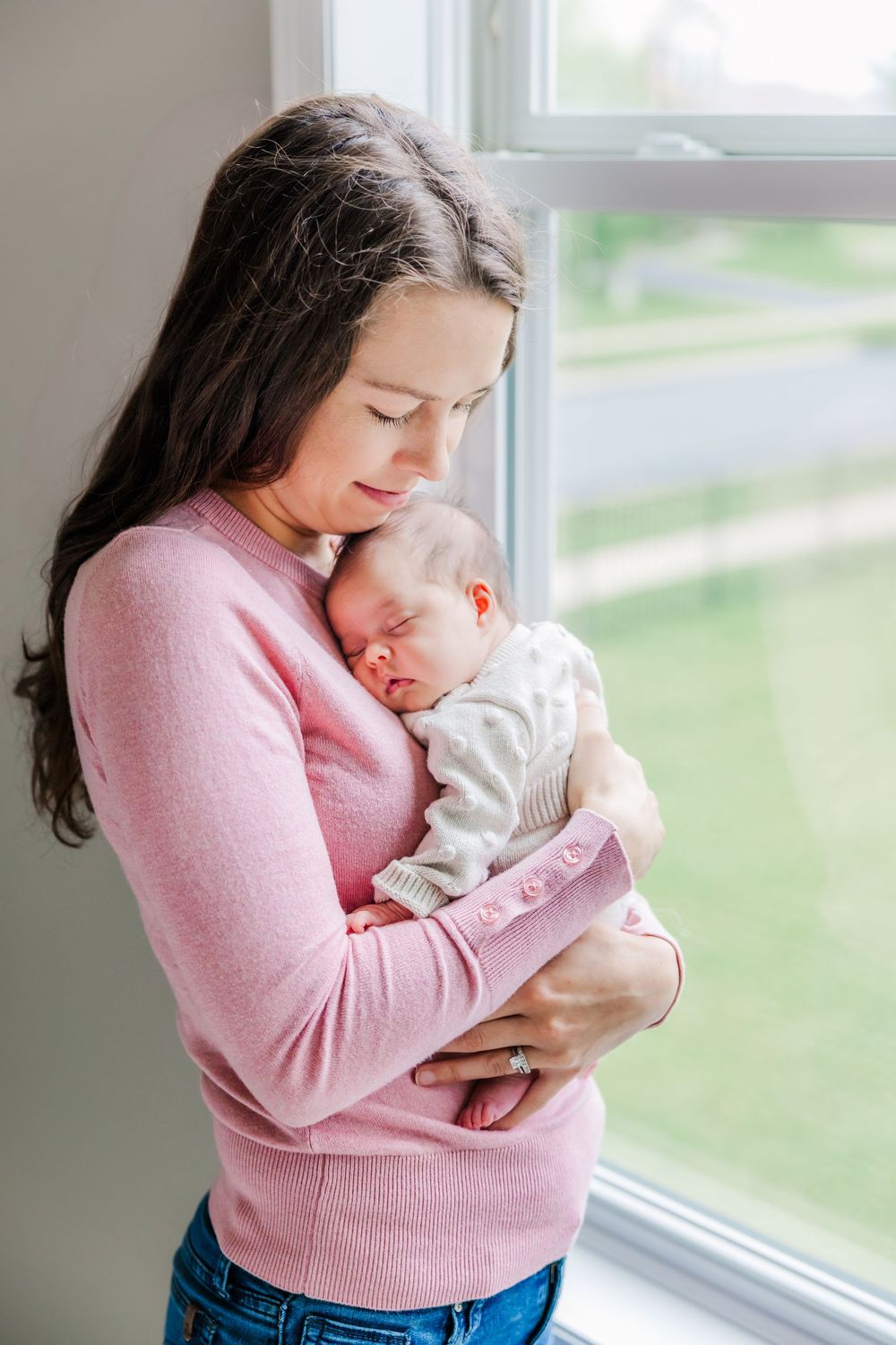 A tender moment between mother in pink sweater holding sleeping newborn baby by a bright window.