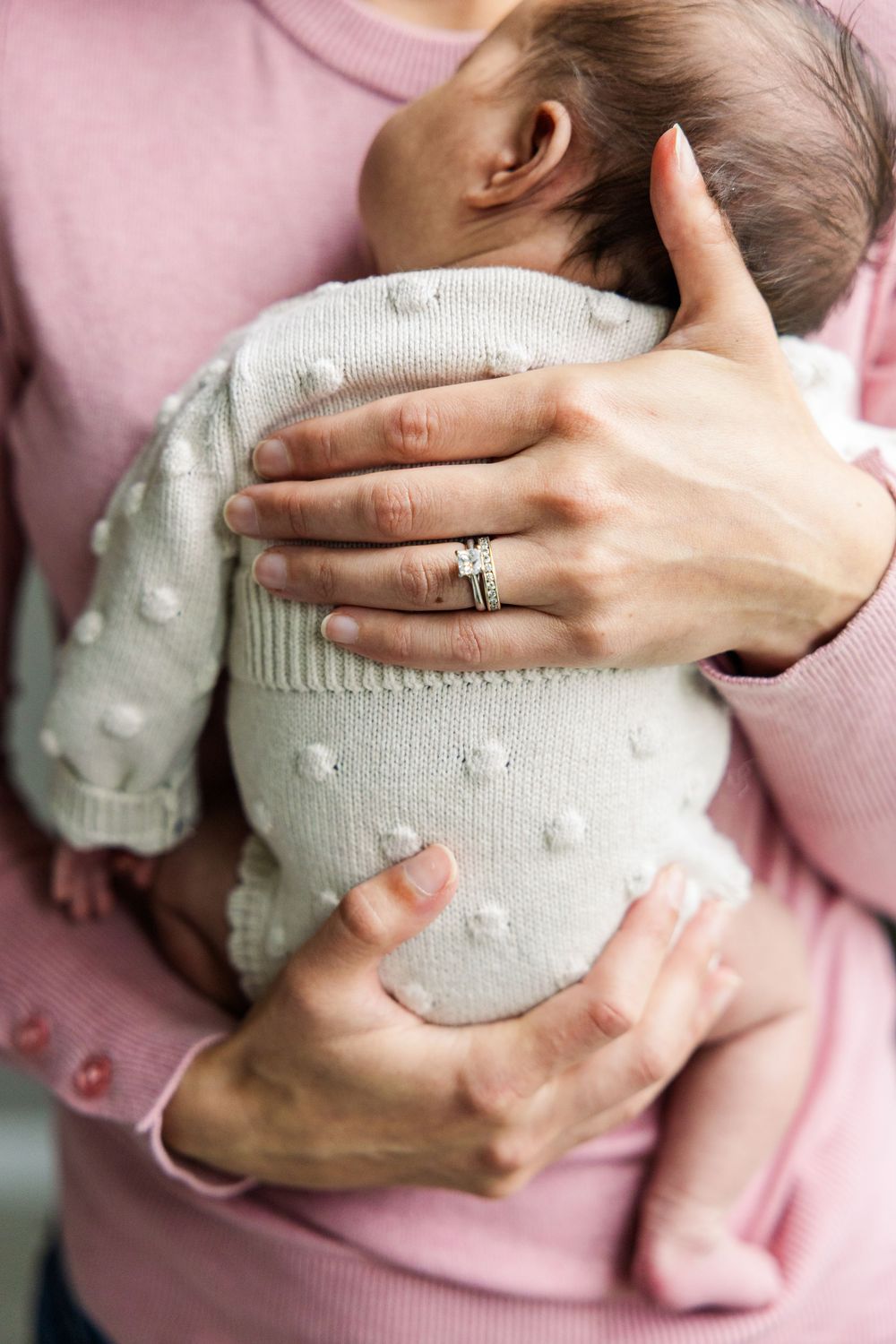 Parent tenderly holds sleeping baby wearing white knitted sweater while wearing soft pink clothing and wedding ring.