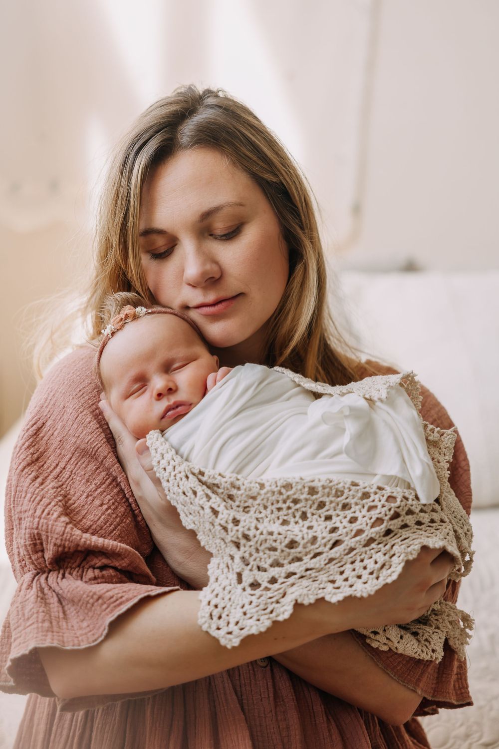 Parent in pink dress holding sleeping newborn wrapped in lace blanket.