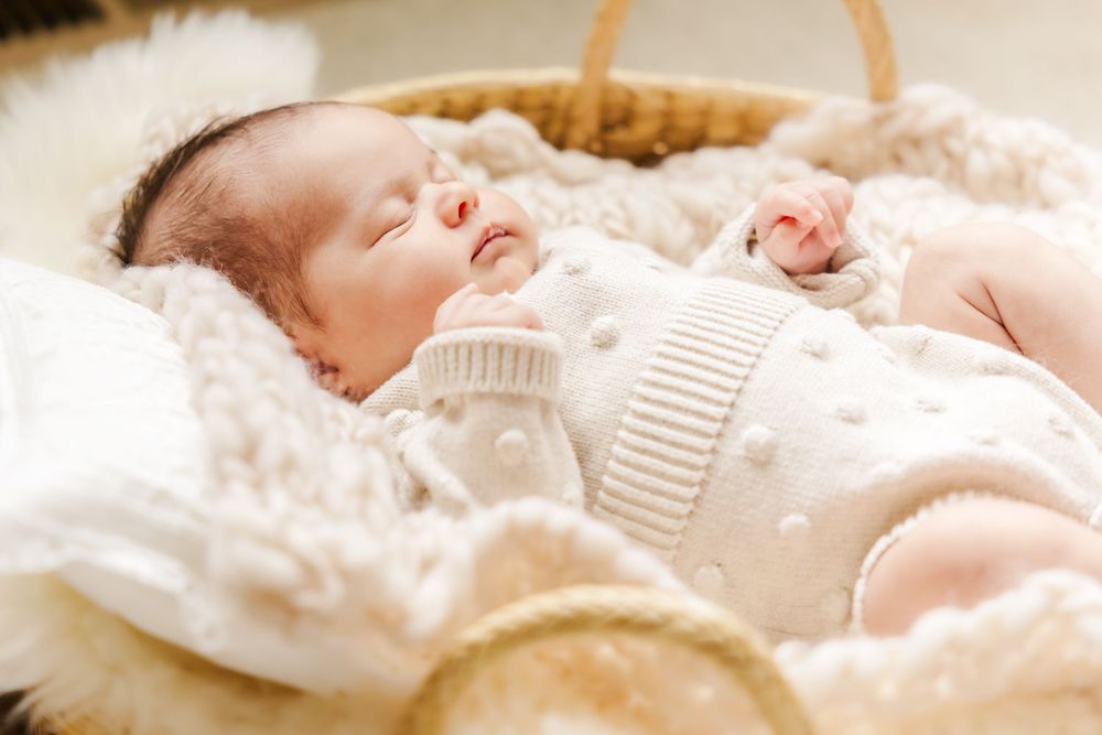 A sleeping baby wearing a white knitted sweater lies peacefully in a basket filled with soft blankets.