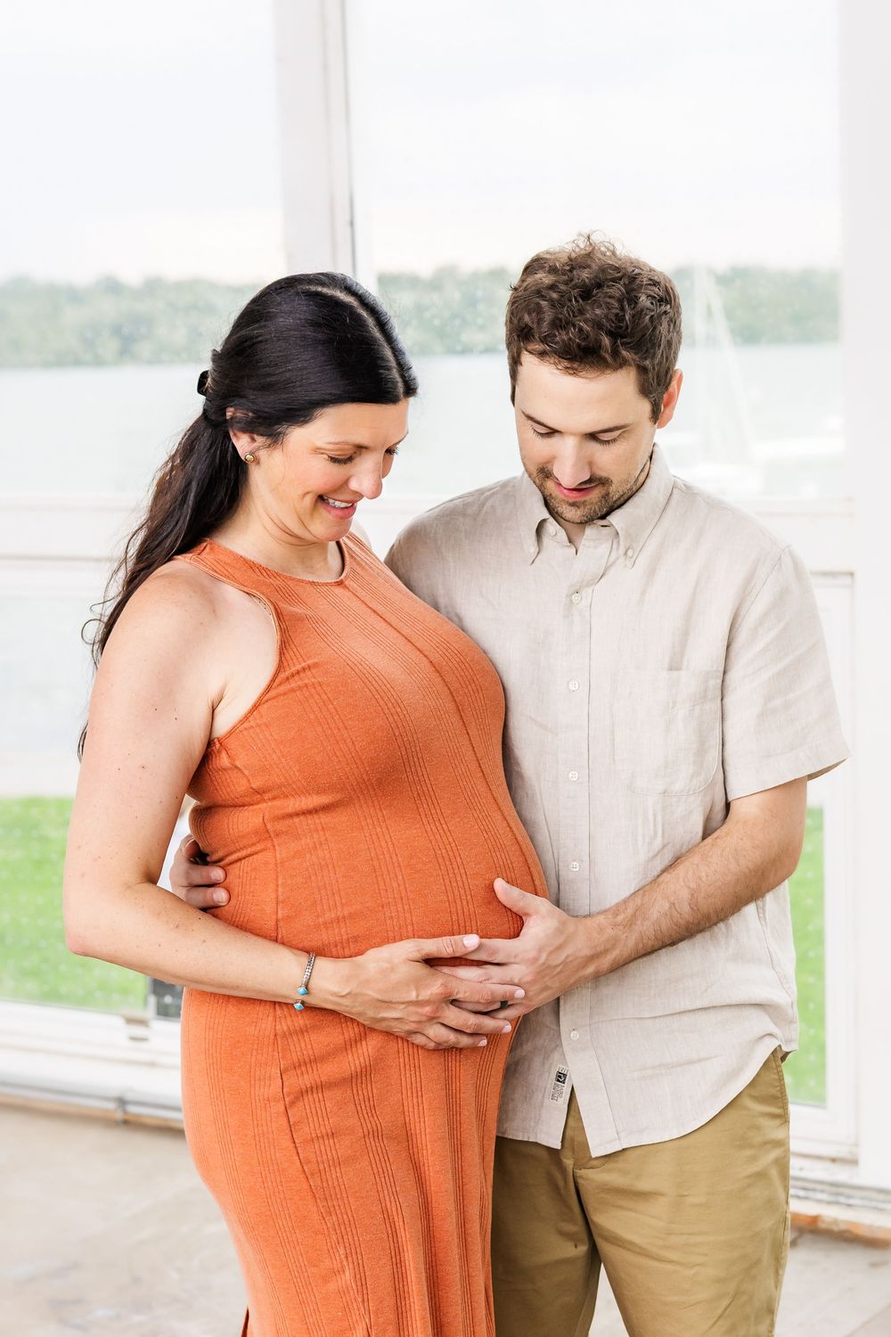 Expecting couple share tender moment together as they touch her pregnant belly wearing orange dress and beige shirt.