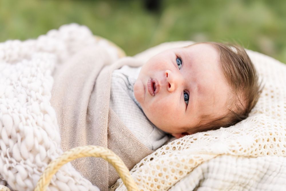 A baby wearing a gray sweater lies on a white textured blanket while looking up with bright eyes.