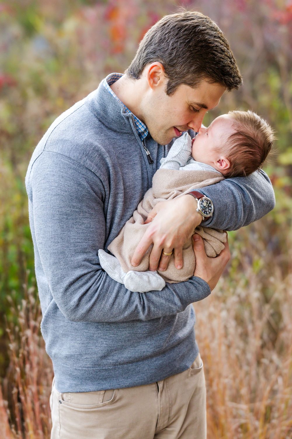 A parent in a gray sweater lovingly cradles and kisses a small baby in white clothing outdoors among autumn foliage.
