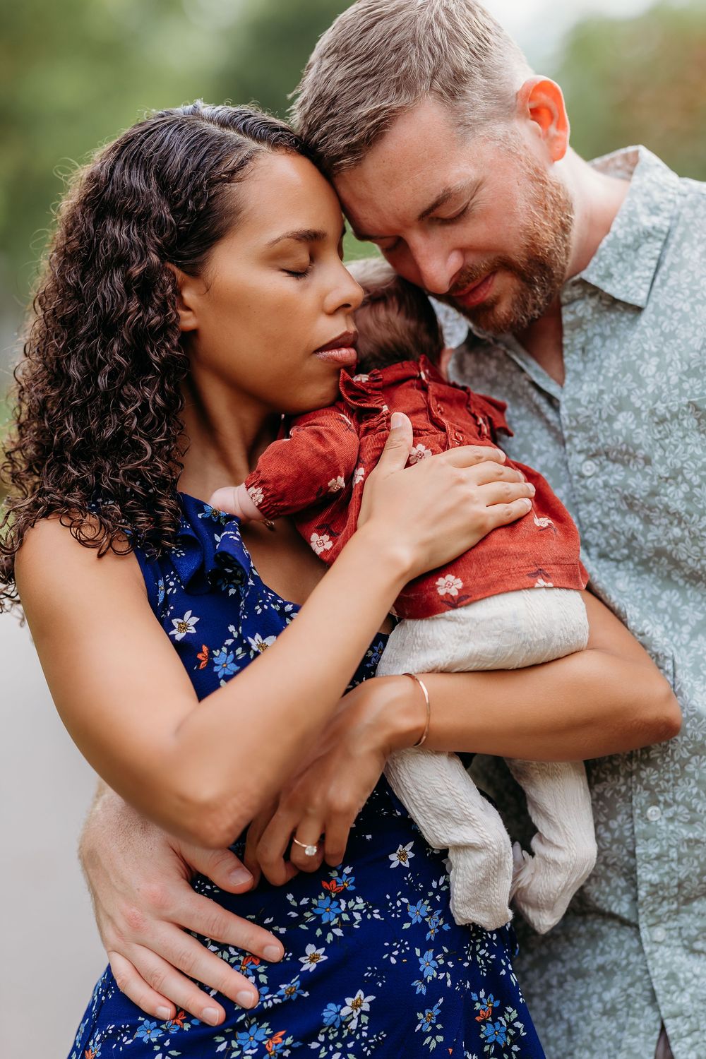 A couple shares an intimate moment while holding their baby wrapped in red clothing outdoors.