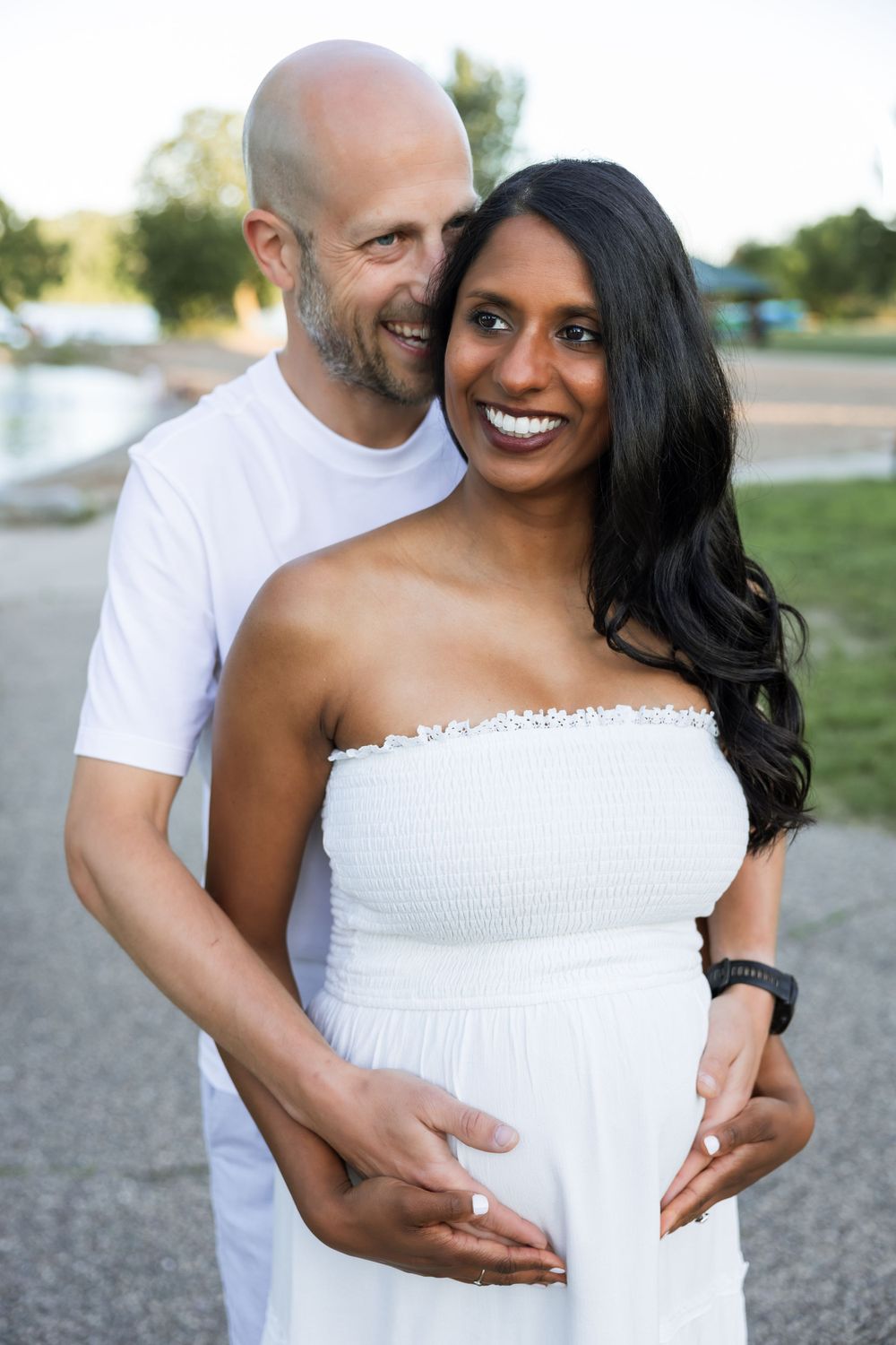 An expectant couple embraces outdoors, with the person in white wearing a strapless dress.