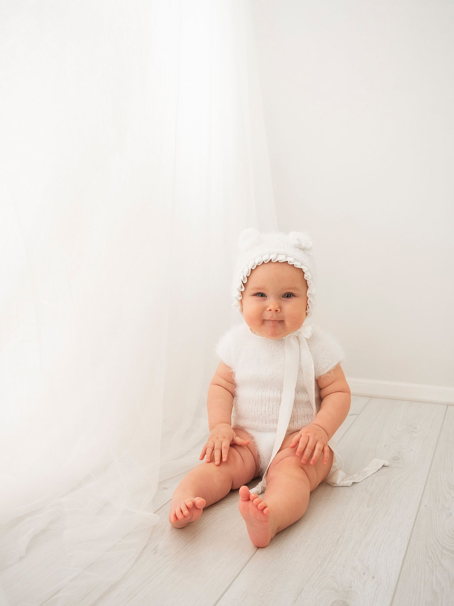 photo of baby girl sitting by curtain, wearing a white fuzzy vest and bonnet with teddy ears.