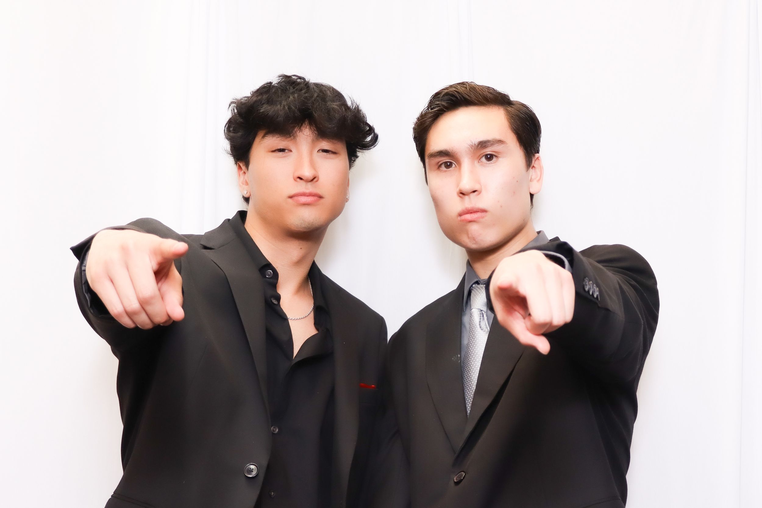 Two men in black suits pointing at camera against white backdrop.