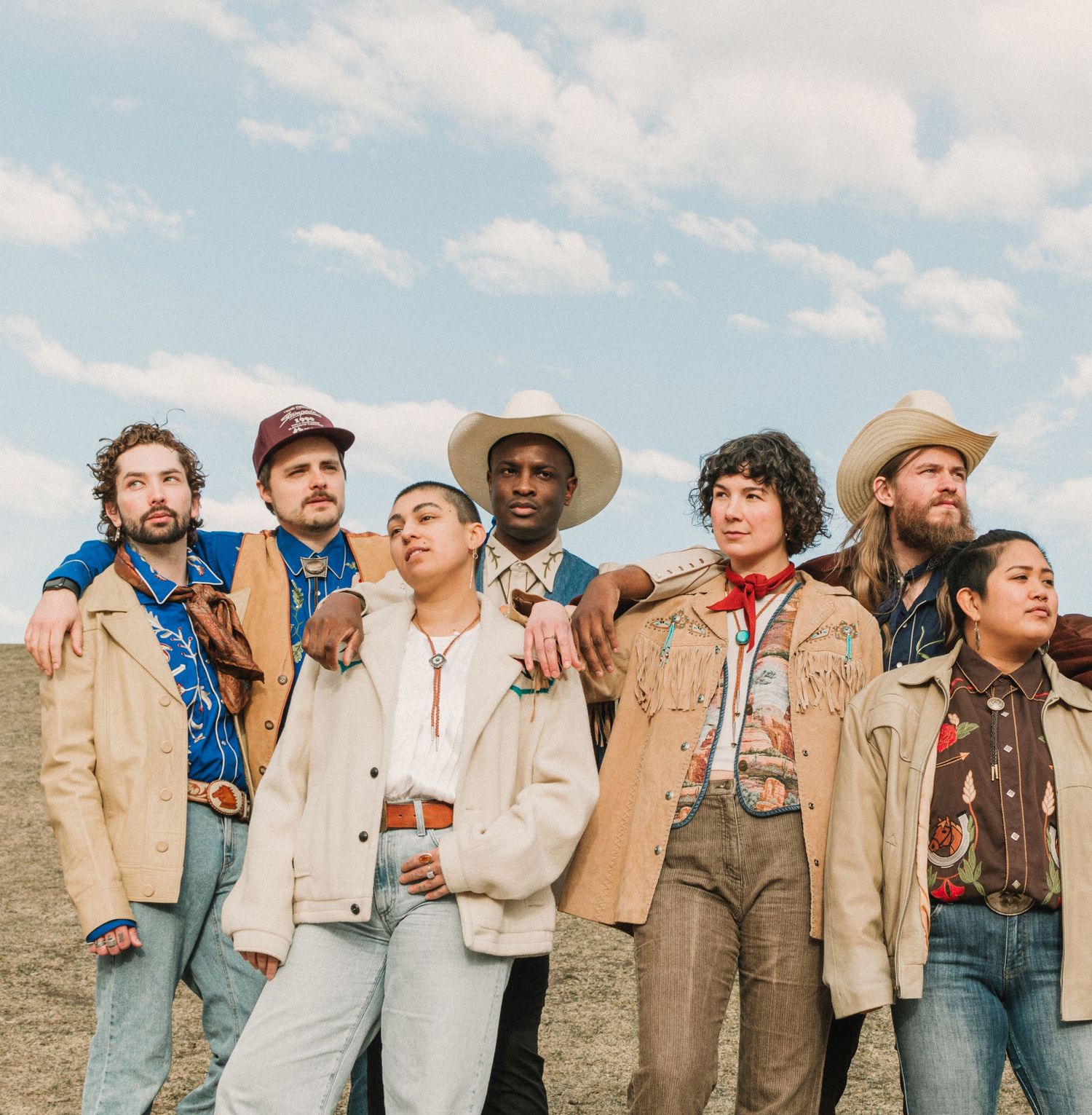 Group of diverse musicians posed together against a blue sky backdrop wearing neutral colored clothing.