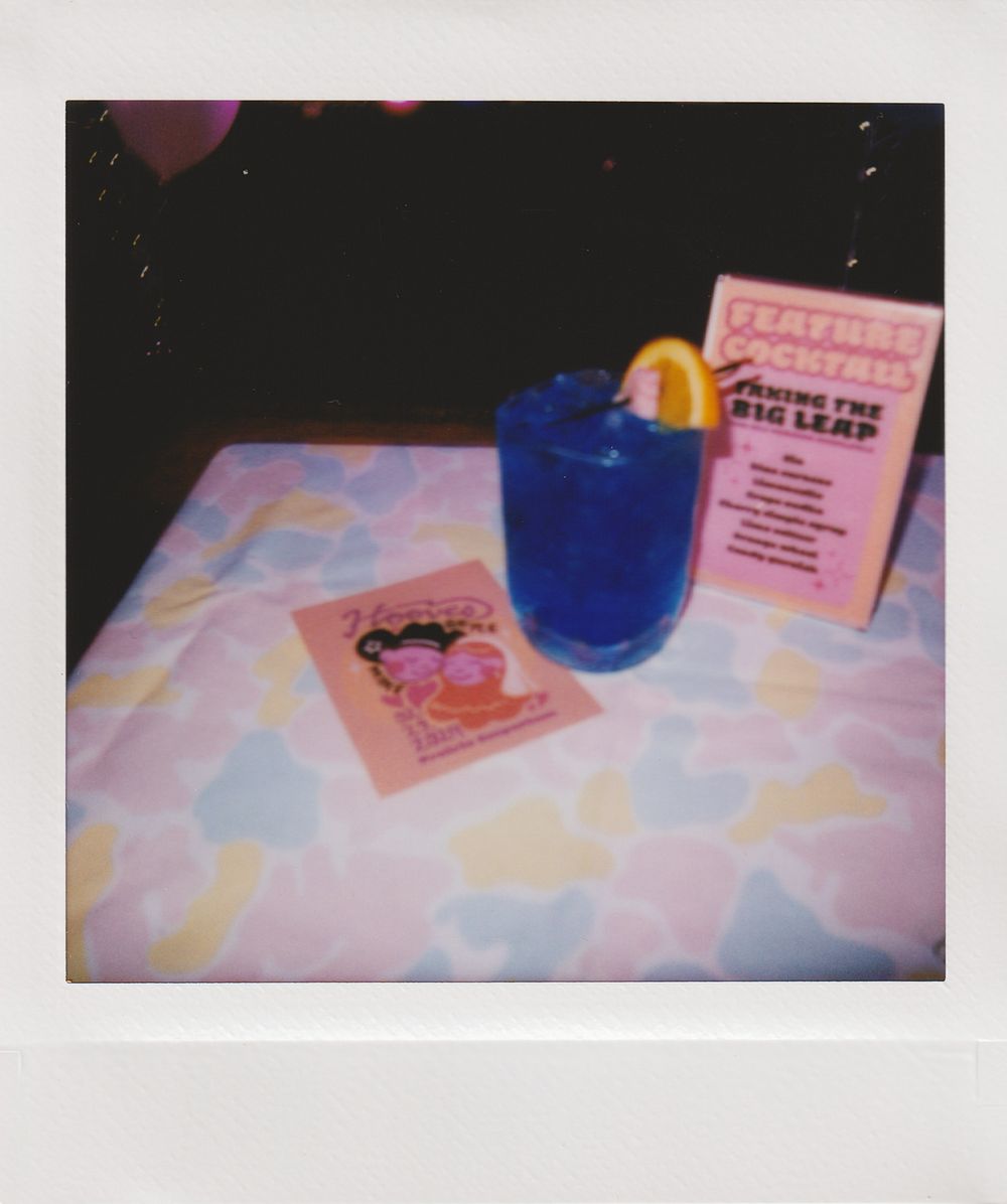 Blue cocktail served with an orange slice on a pastel patterned tablecloth next to a menu and pink card.