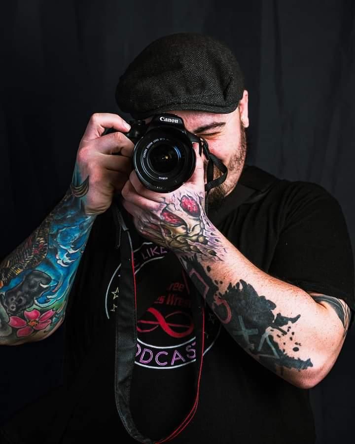 Photographer with colorful tattoo sleeves taking picture with Canon camera against black background.