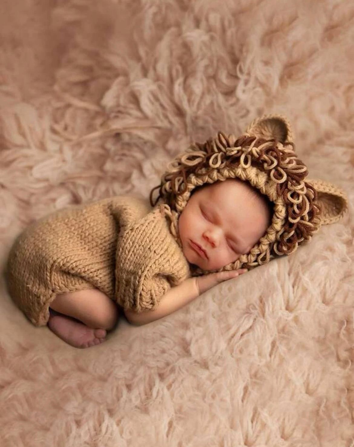 Newborn baby sleeping peacefully in brown knit outfit on textured beige backdrop.
