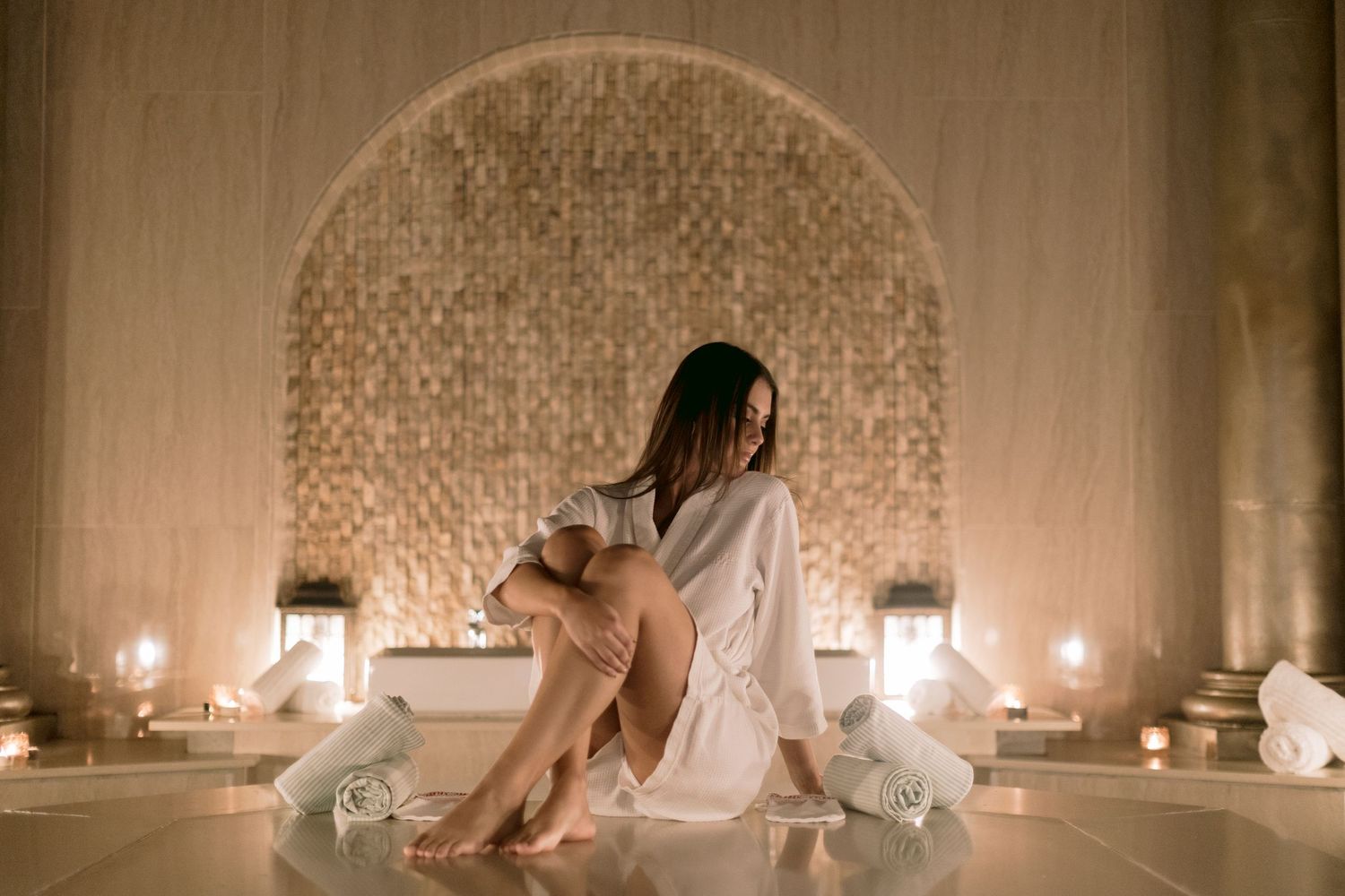 Woman in white robe relaxing in luxurious candlelit spa hammam with mosaic tile arch backdrop.