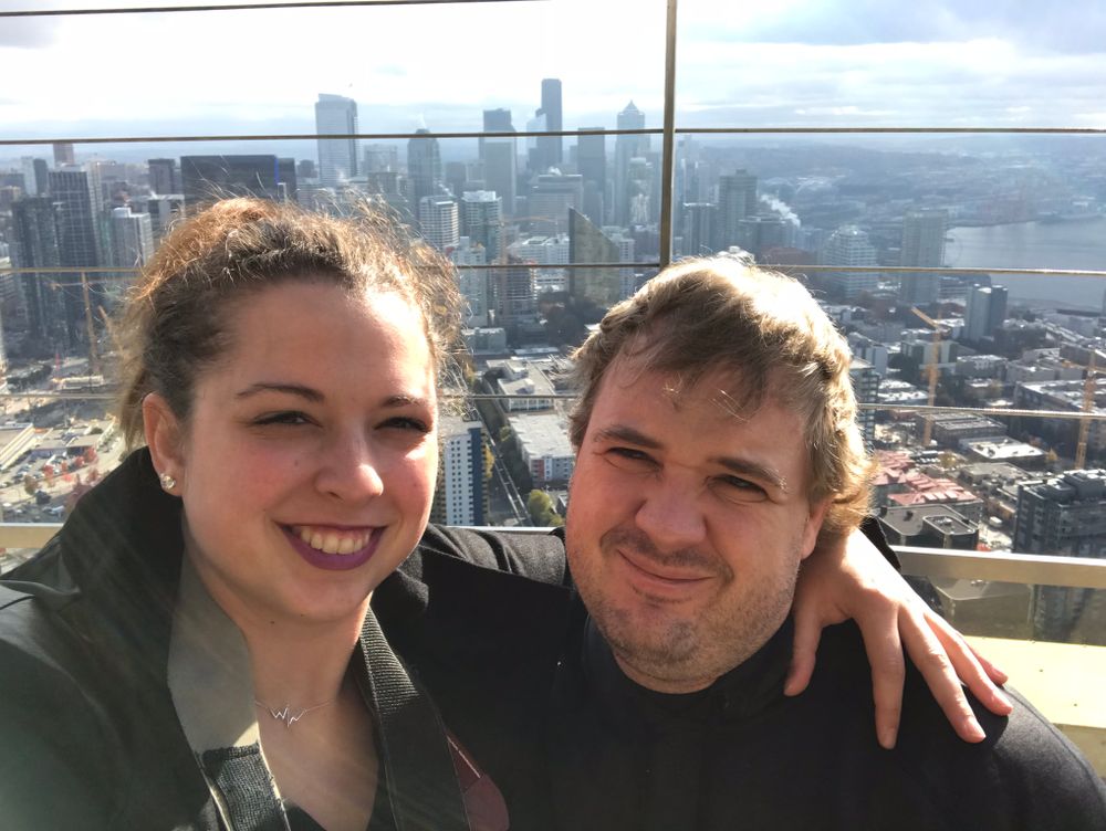 Two people posing for a selfie with a metropolitan skyline in the background.