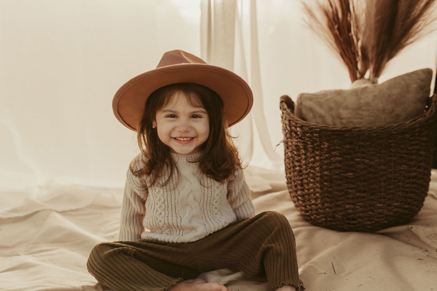 Child wearing brown hat and sweater sits cross-legged by woven basket with pampas grass.