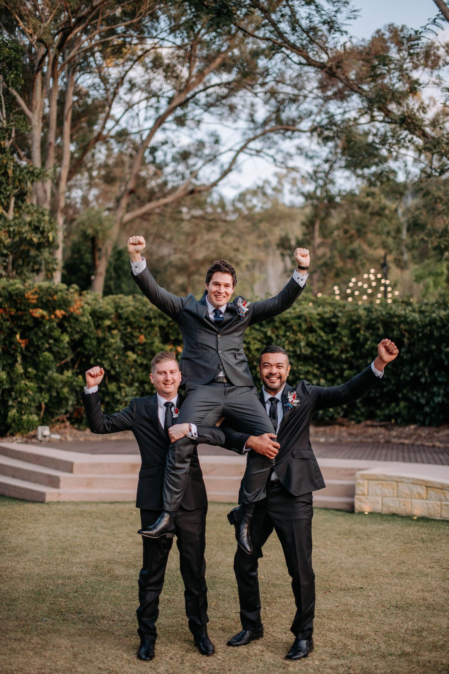 Groomsmen lift and celebrate with joyful expressions on manicured lawn.