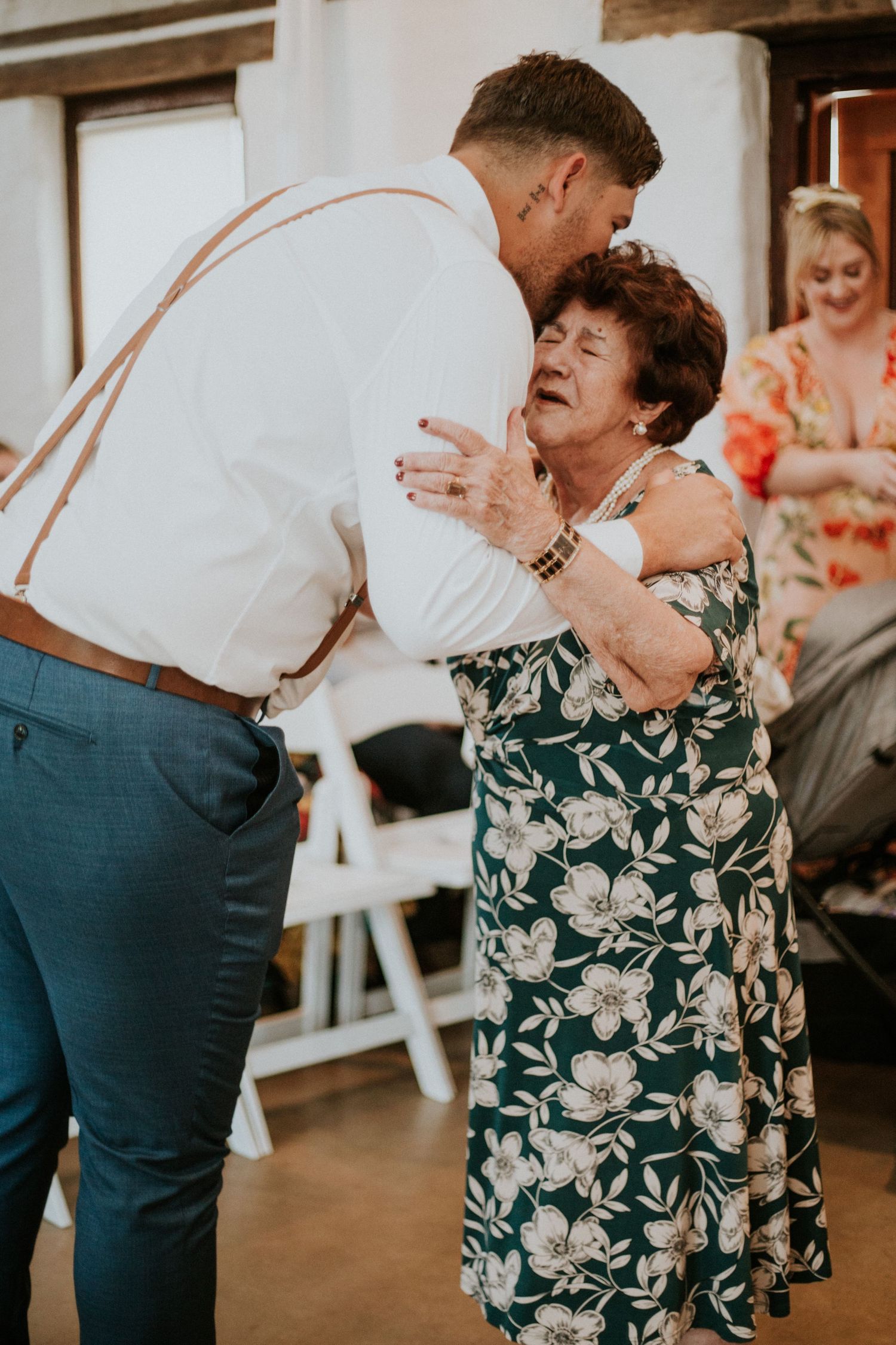 Groom in white shirt and suspenders sharing a warm dance with elderly woman at wedding reception.