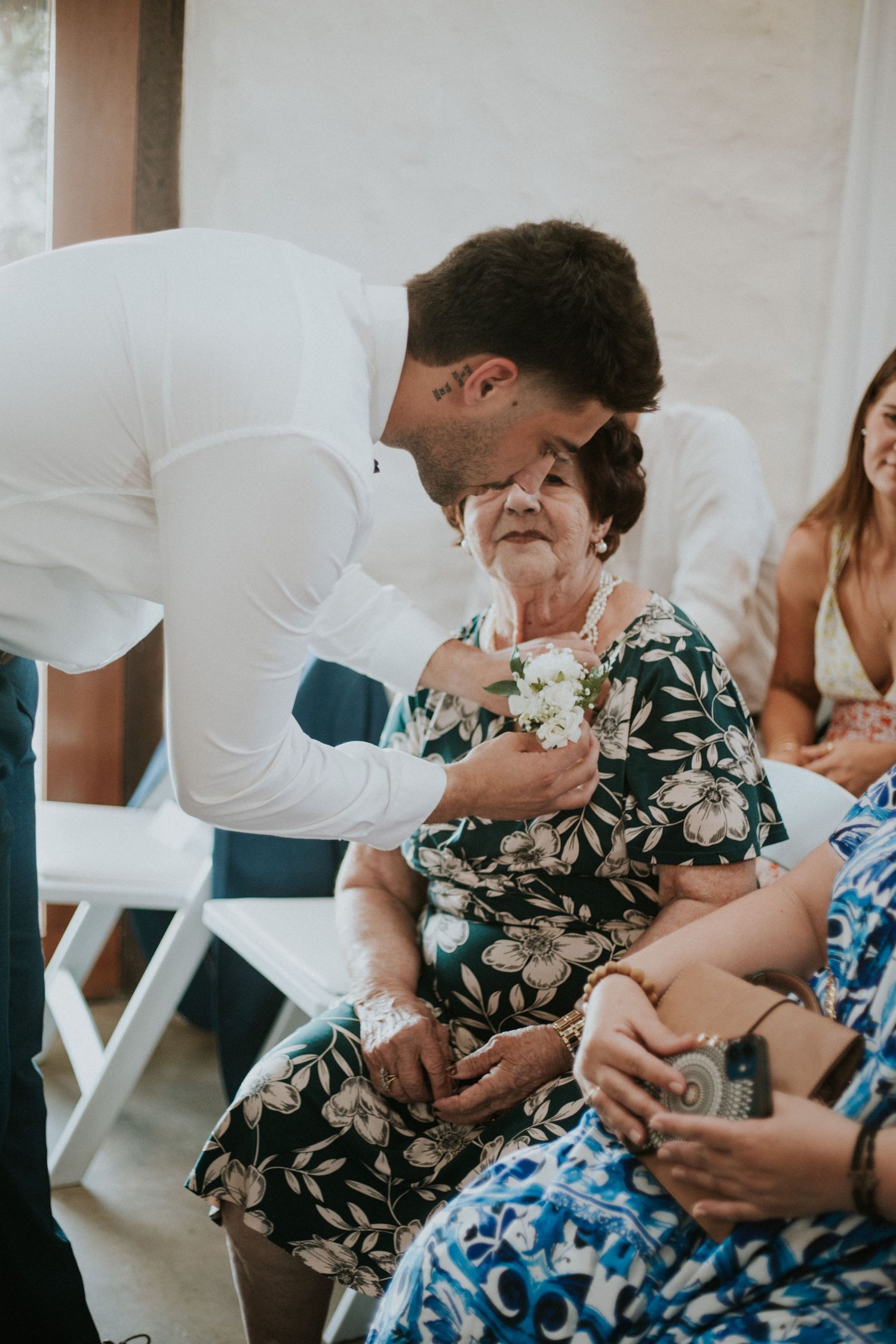 Groom in white shirt affectionately kissing elderly woman in floral dress seated at wedding ceremony.