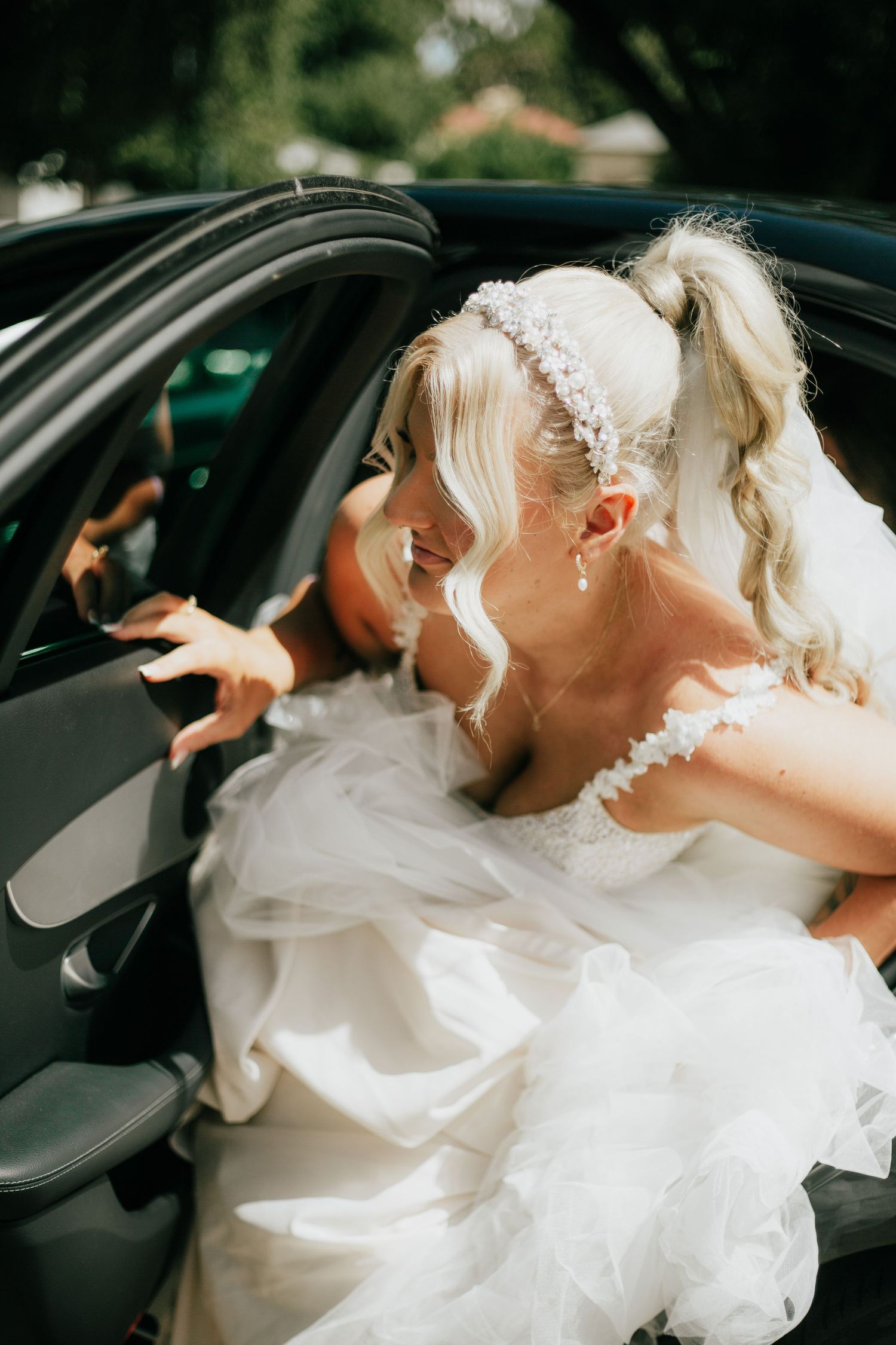 Smiling bride in white ruffled gown and floral headband stepping out of a black wedding car.