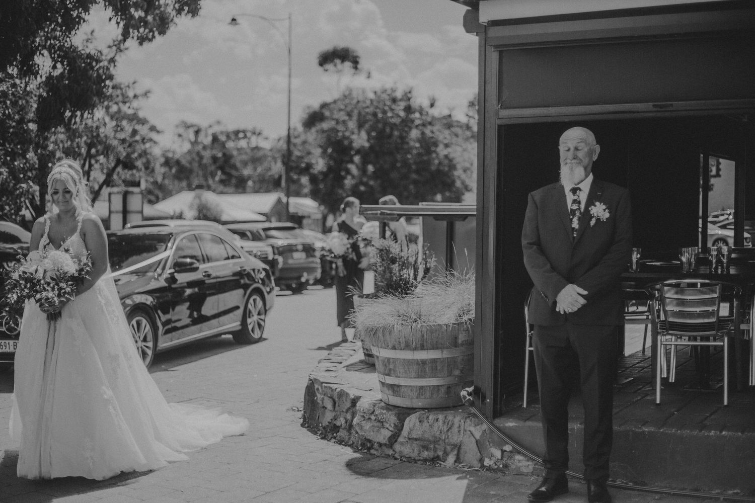 Black and white photo of bride in gown approaching man with boutonniere outside rustic wedding venue.