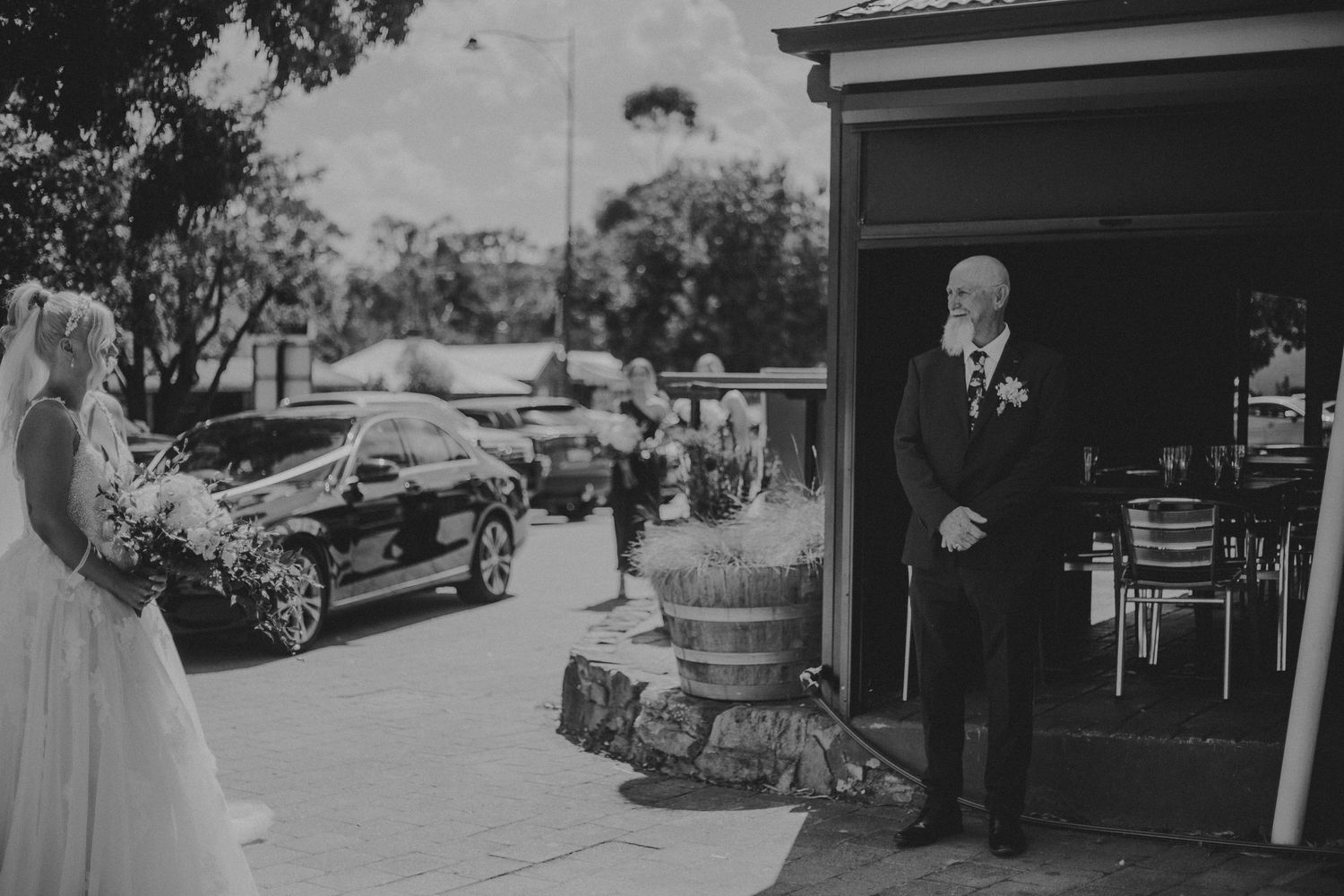 Black and white photo of bride holding bouquet facing older man in suit outside rustic wedding venue.
