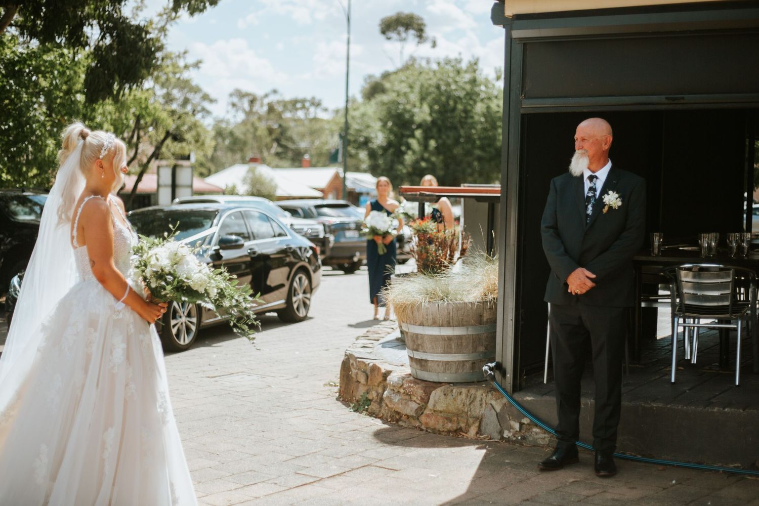Bride in white gown holding bouquet shares emotional first look moment with older man outside venue.