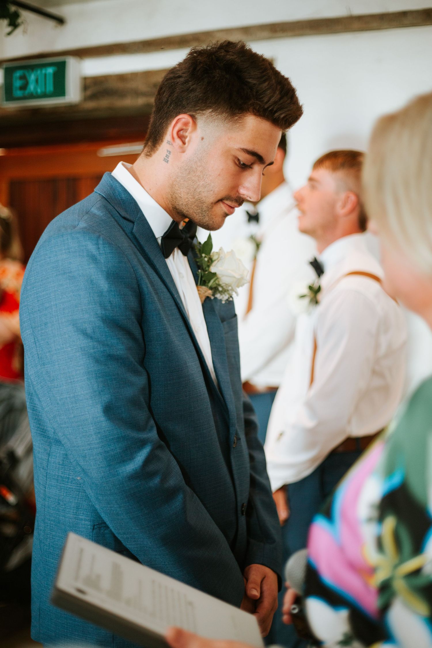 Groom in blue suit and bow tie reads during indoor wedding ceremony with guests nearby.