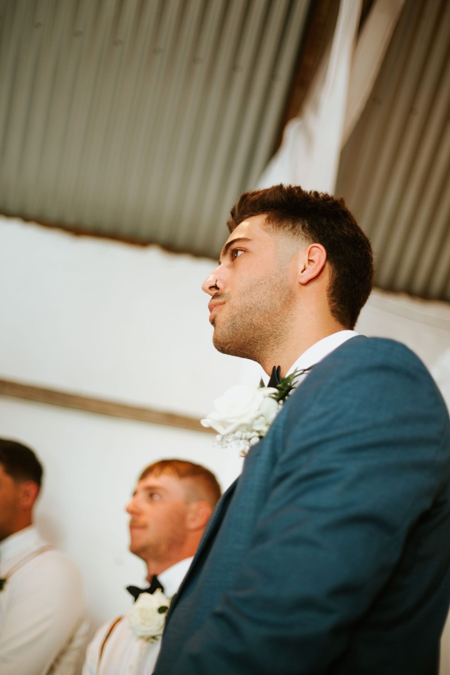 Groom in blue suit with white boutonniere looks up during wedding ceremony in rustic venue.