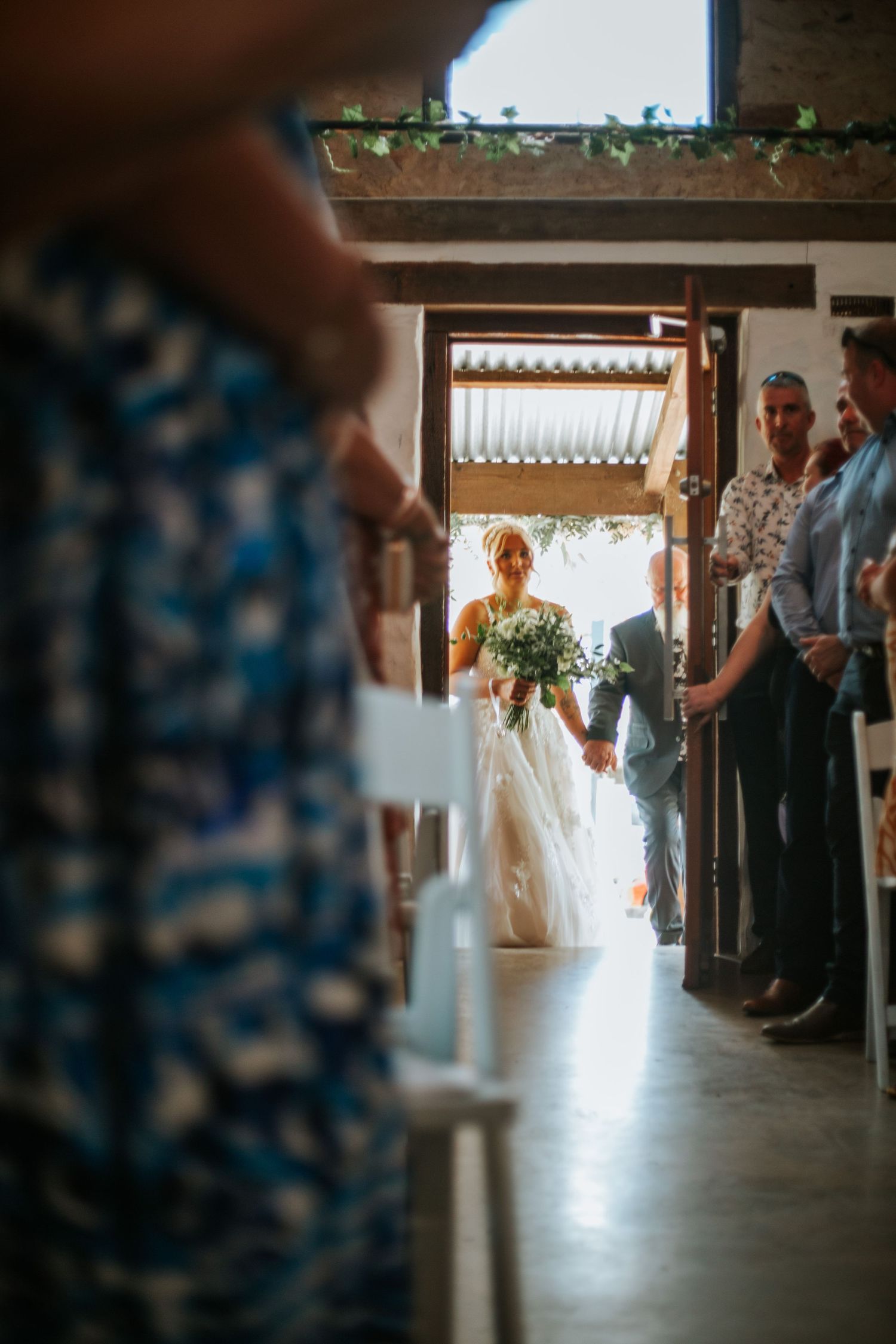 Bride in white lace gown walks down aisle with escort through ceremony venue doorway.
