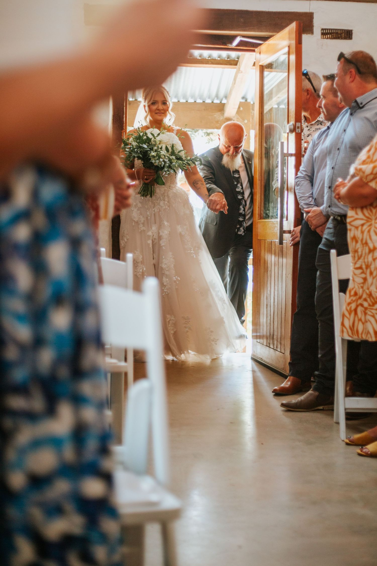 Bride in white lace gown enters wedding ceremony venue escorted down the aisle by family.