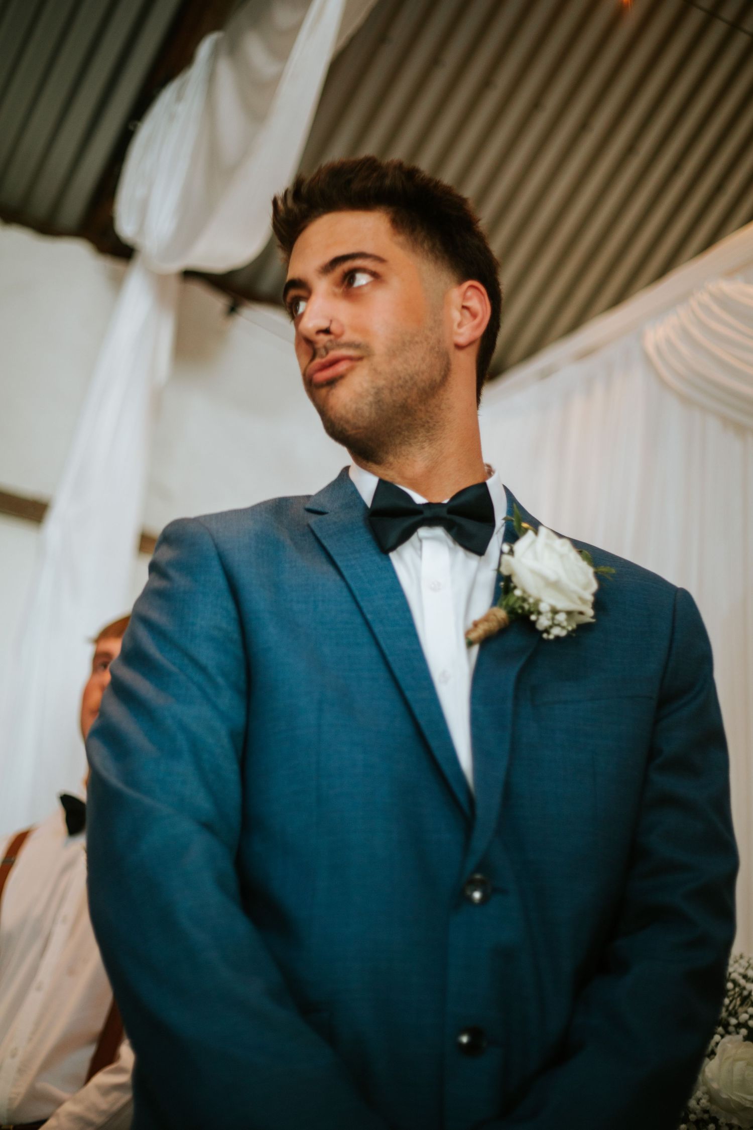 Handsome groom in blue suit and black bow tie watches bride walk down the aisle.