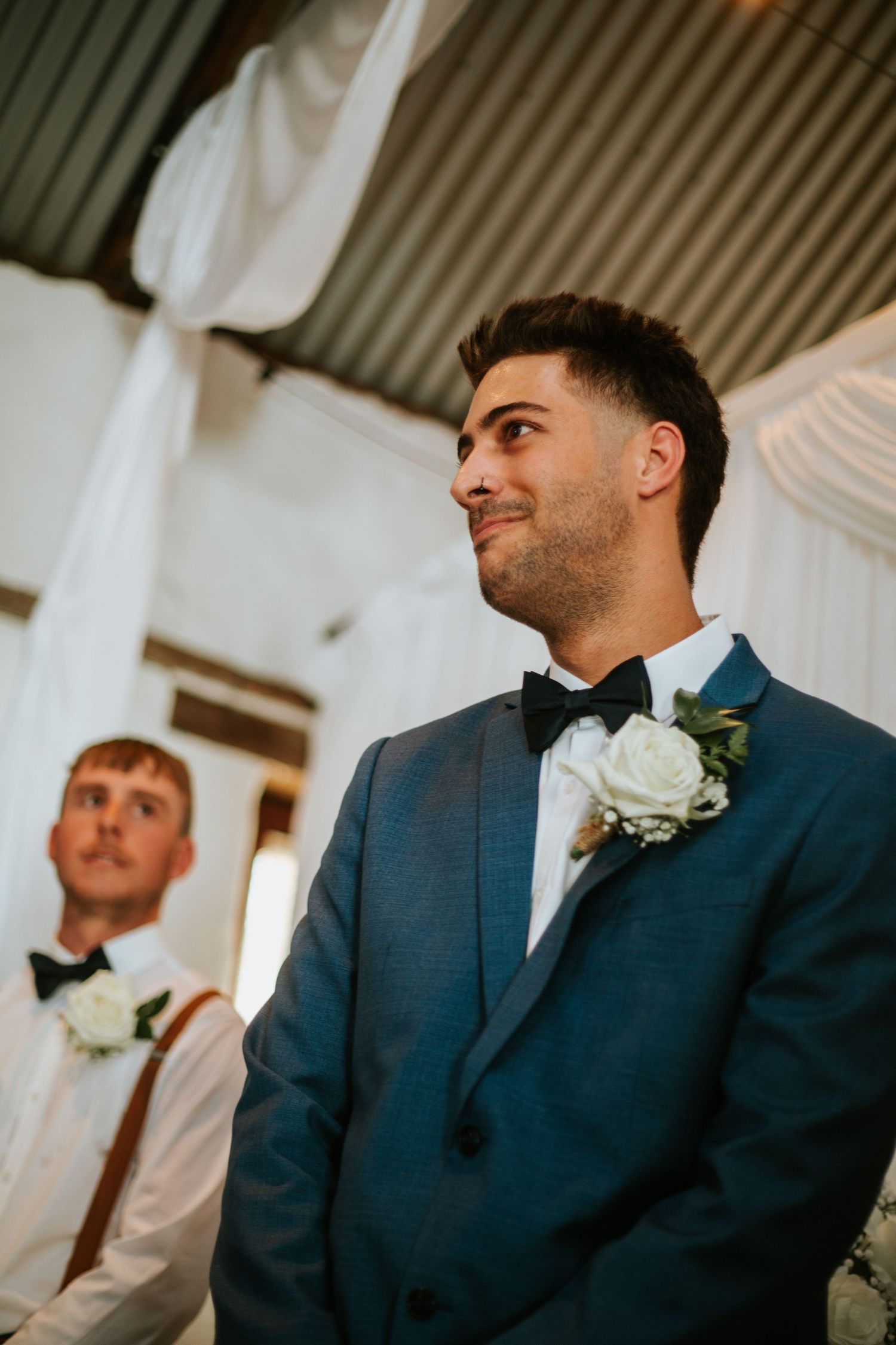 Groom in blue suit smiles with tears as he watches bride approach during wedding ceremony.