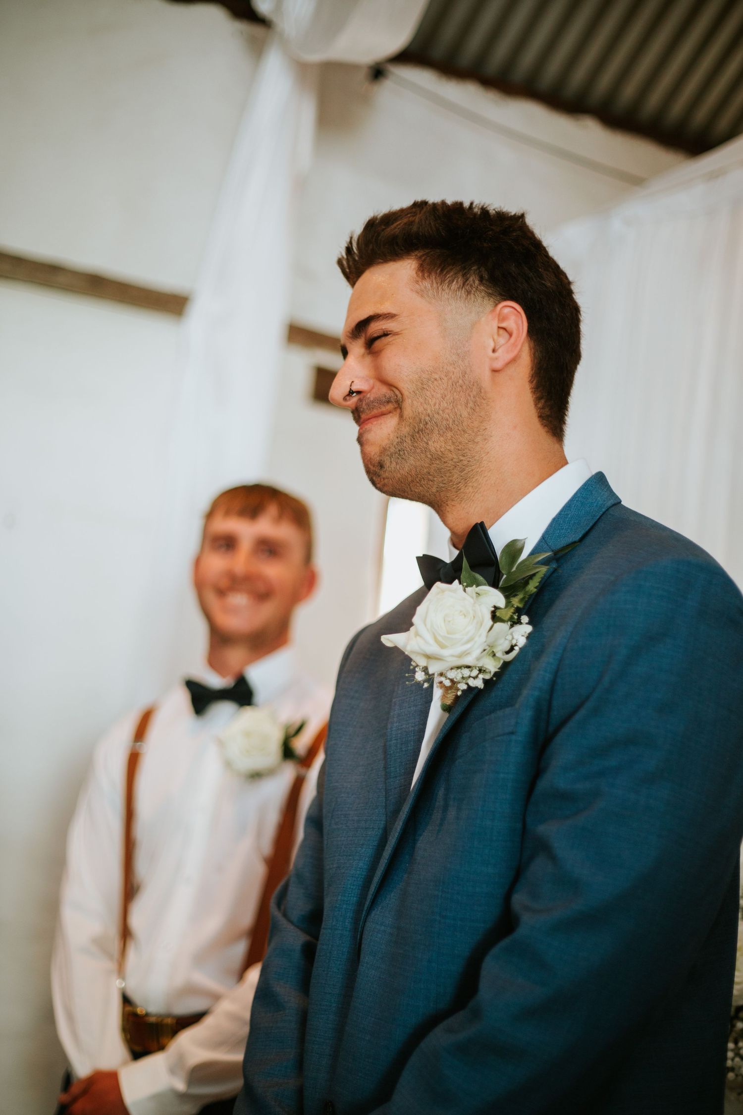Groom in blue suit with white boutonniere smiling at altar during wedding ceremony.