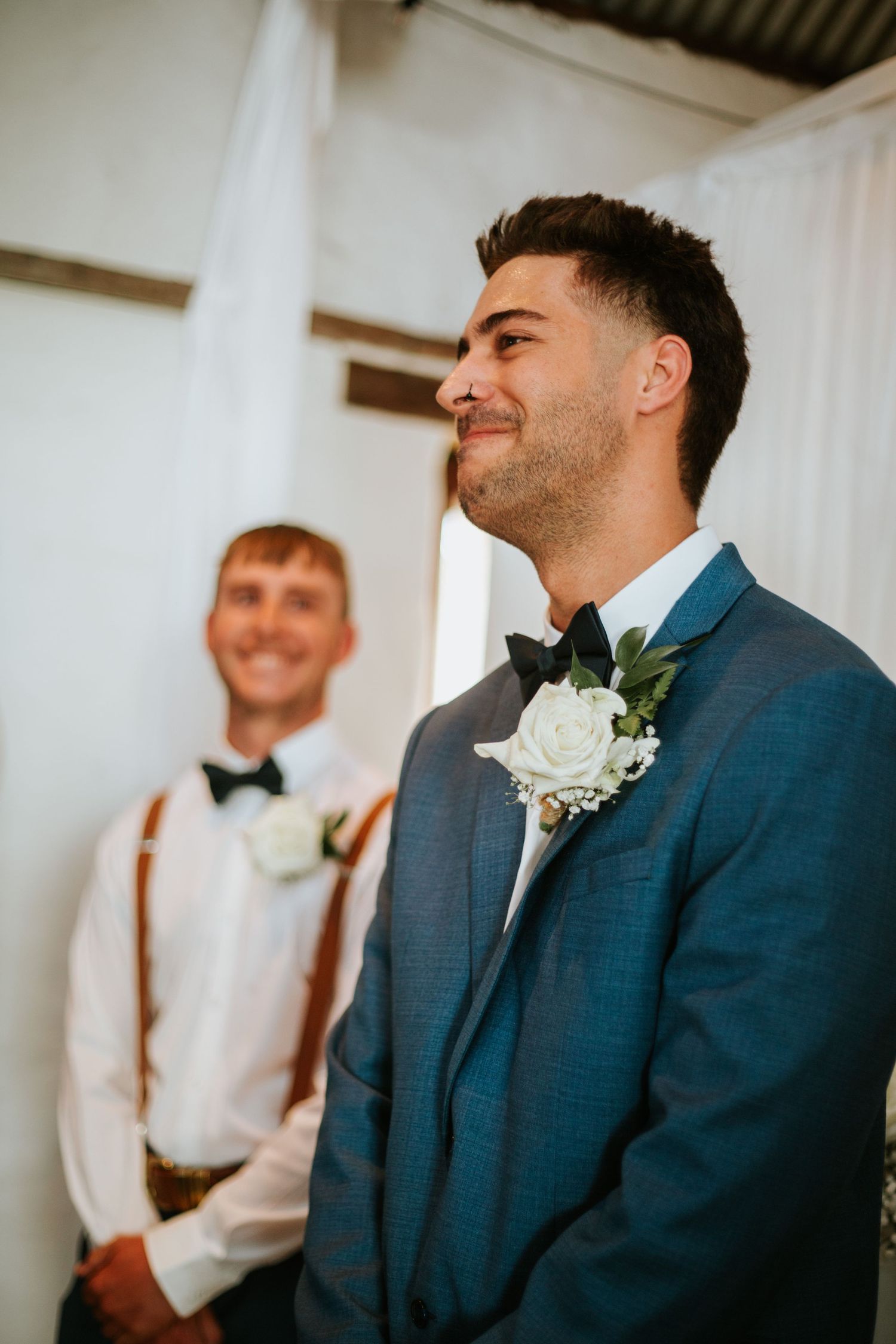 Smiling groom in blue suit and bow tie watching bride walk down the aisle.