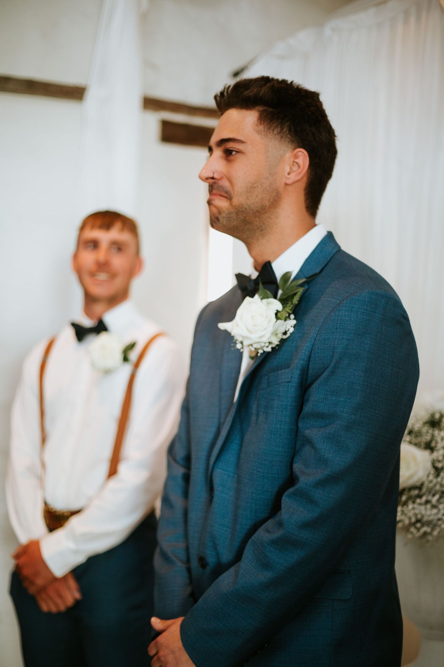 Groom in blue suit with white boutonniere standing at altar with groomsman behind.
