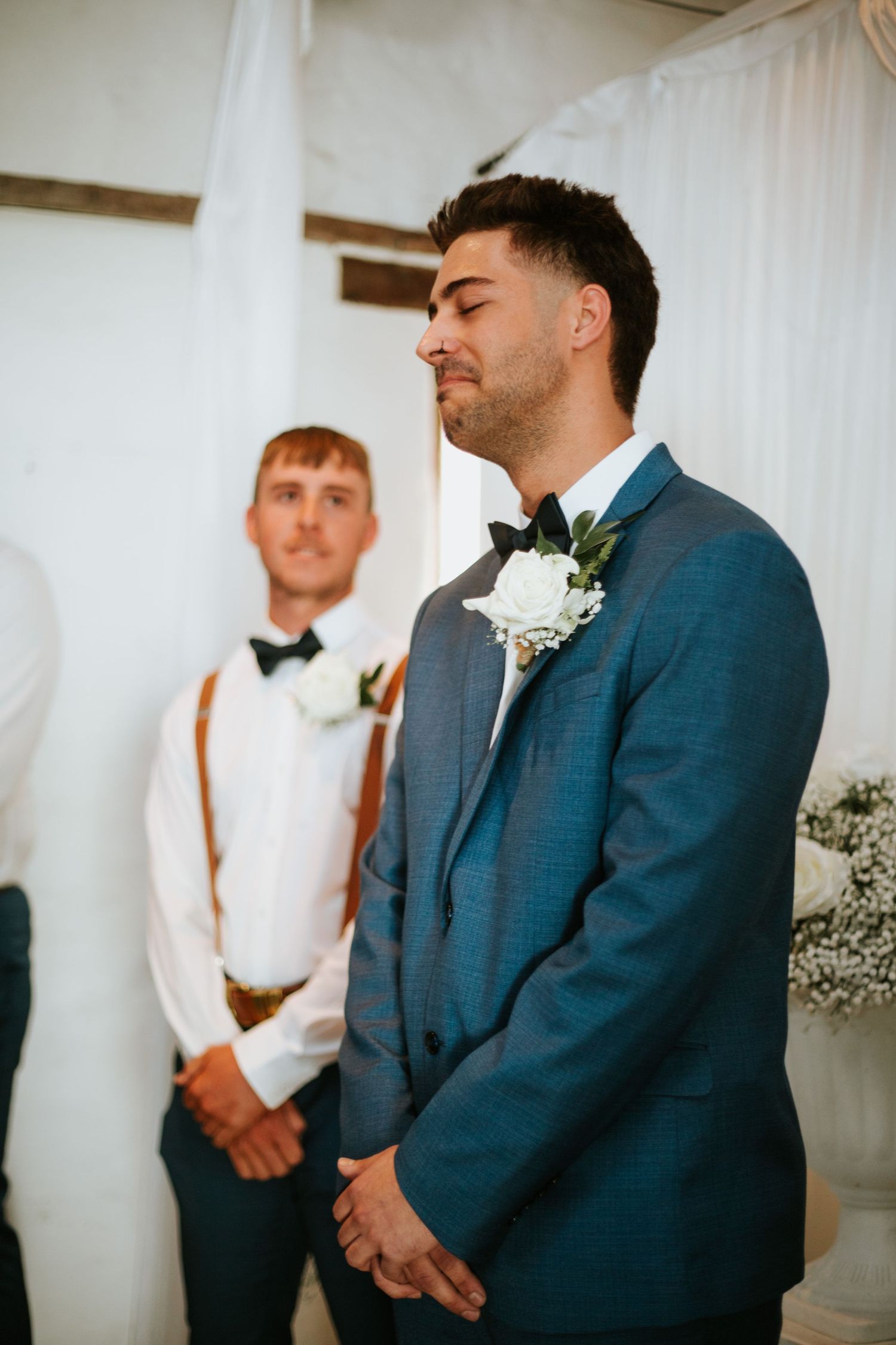 Groom in blue suit with eyes closed, emotional moment at indoor wedding ceremony.